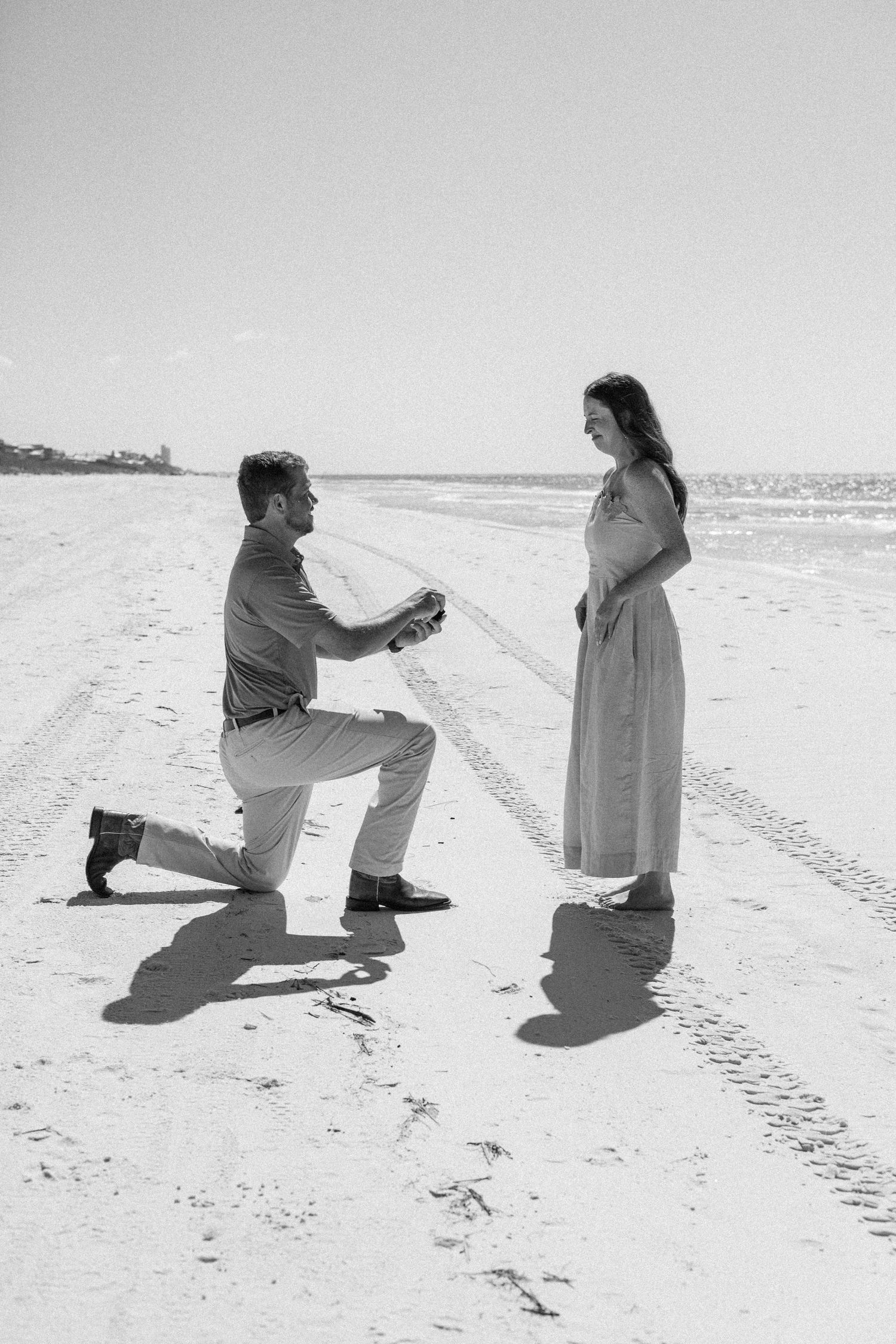 Man proposes on bended knee to woman on sandy beach in romantic black and white photography.