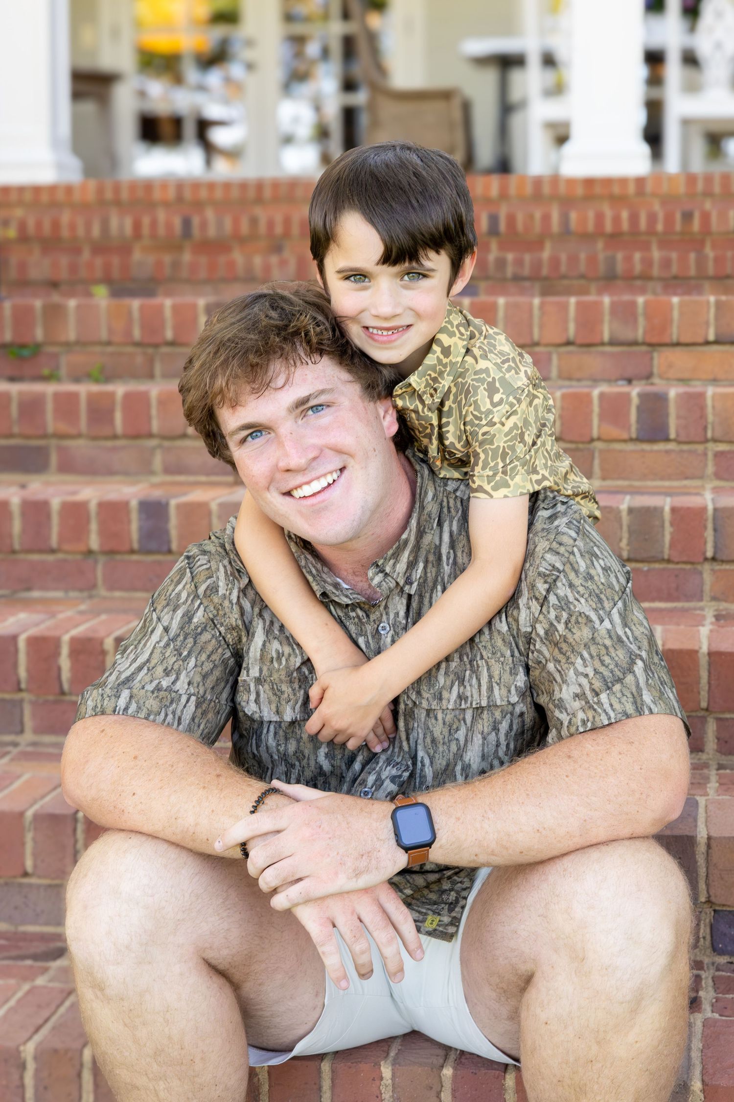 A smiling man and young boy in camo shirts sit together on brick steps, sharing a warm embrace outdoors.