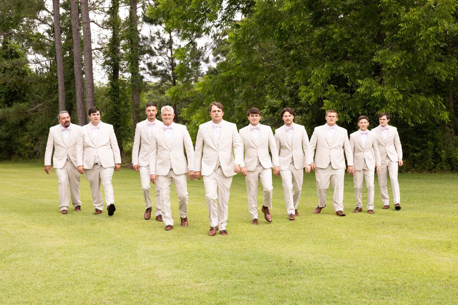 Groom and groomsmen in matching tan suits walking confidently across a green lawn with trees in background.