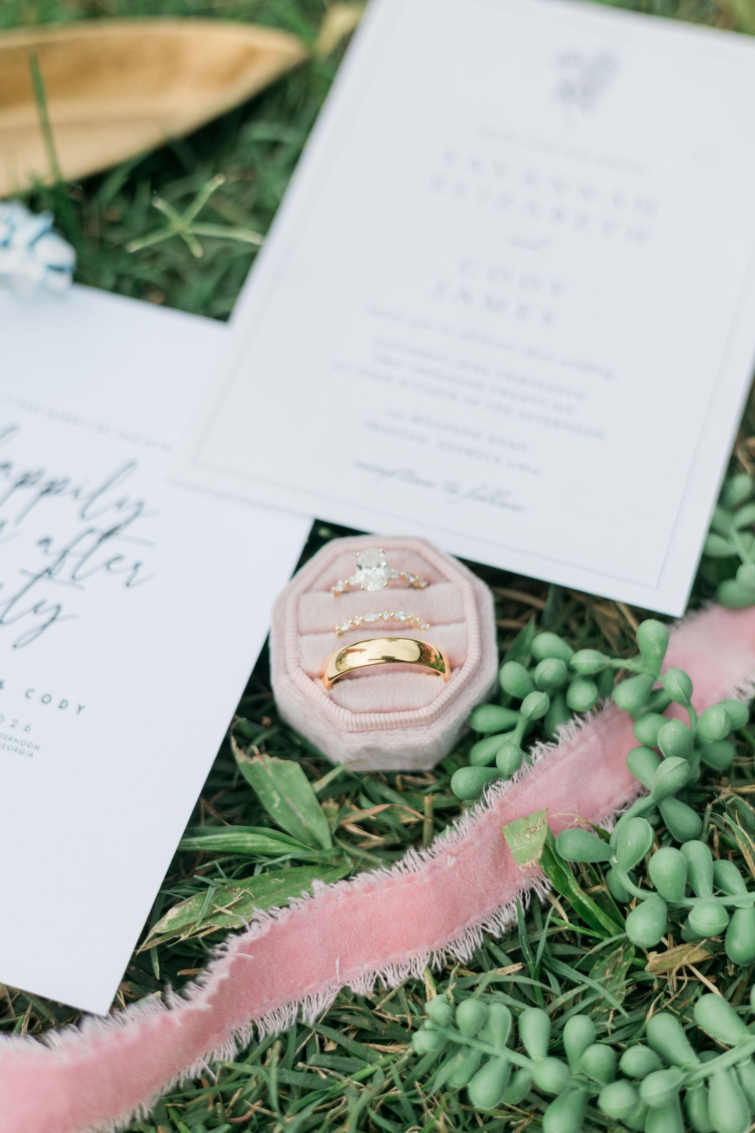 Gold wedding rings in pink velvet box beside wedding invitation suite on green grass.