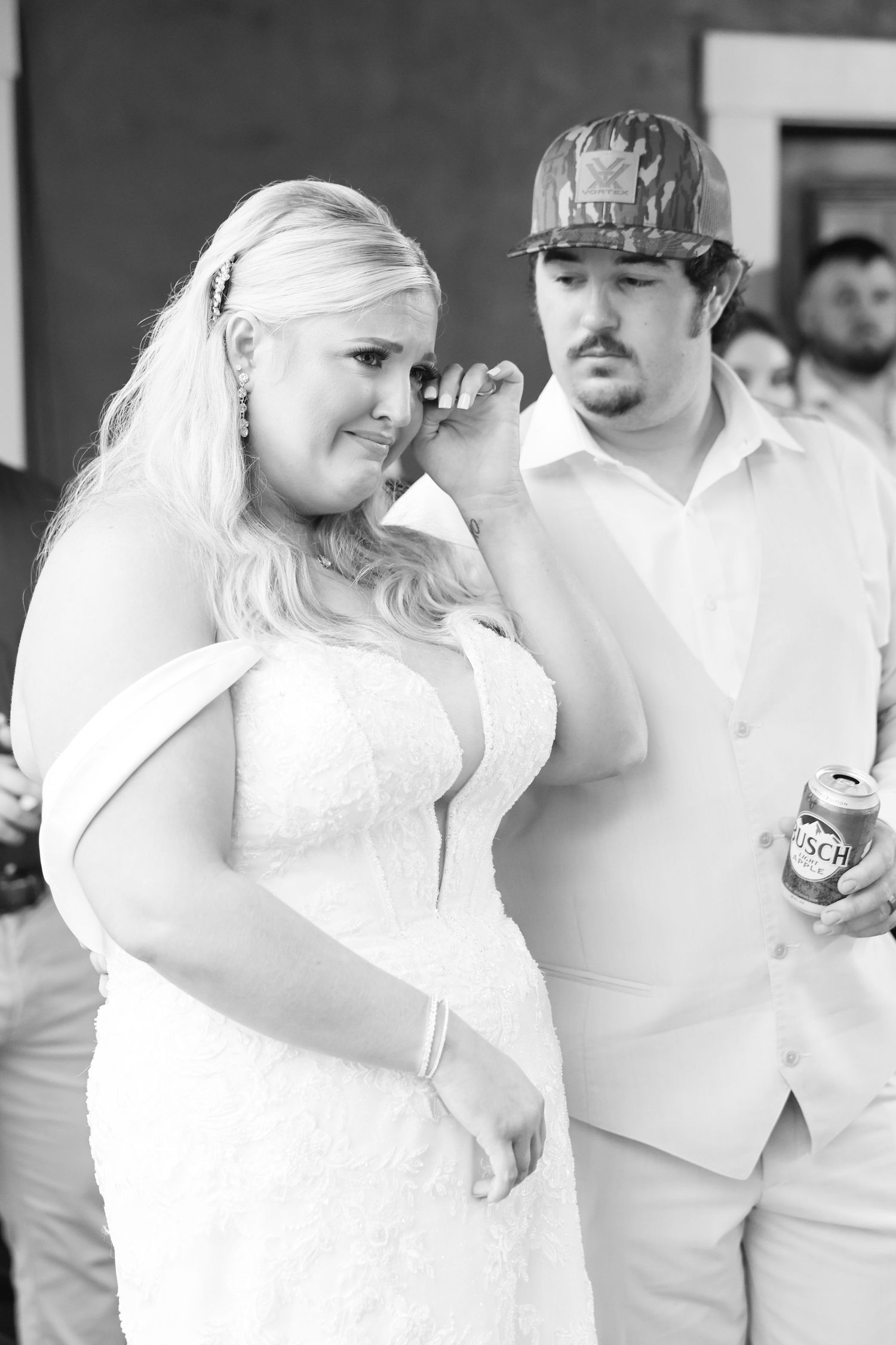 Emotional bride wiping tears beside groom holding beer can at rustic wedding reception, black and white photo.