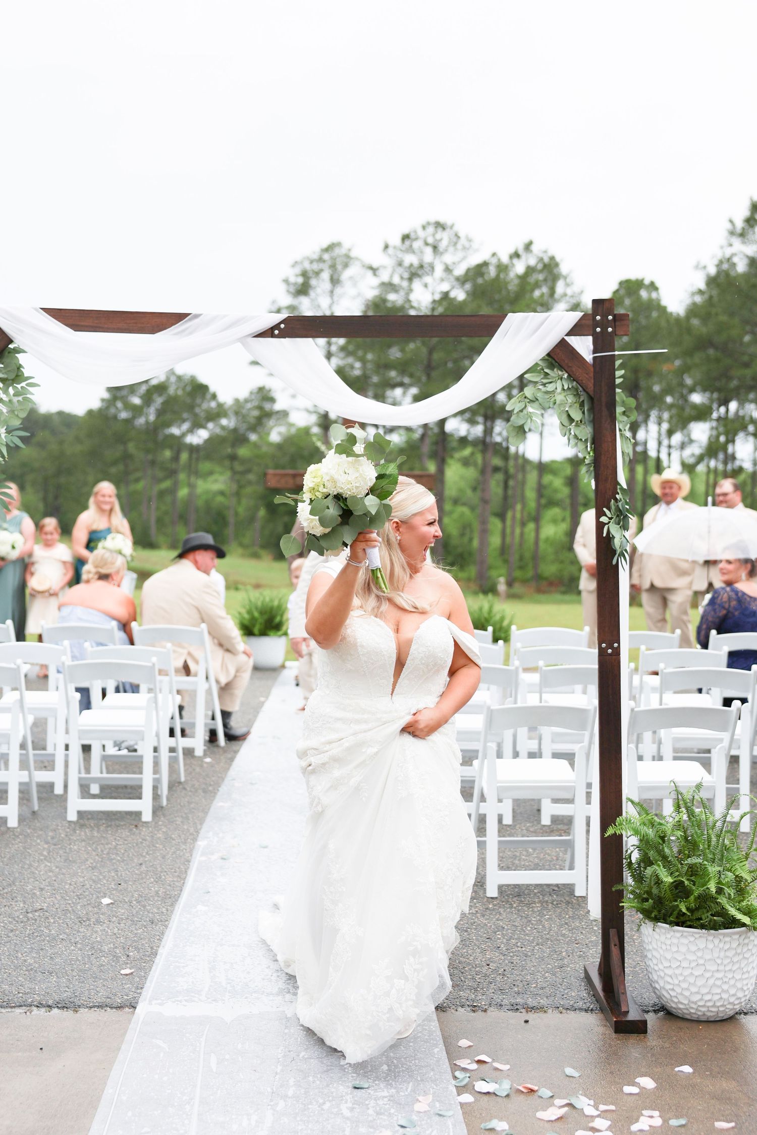 Bride in white lace gown walks down outdoor aisle holding white bouquet at elegant garden wedding ceremony.