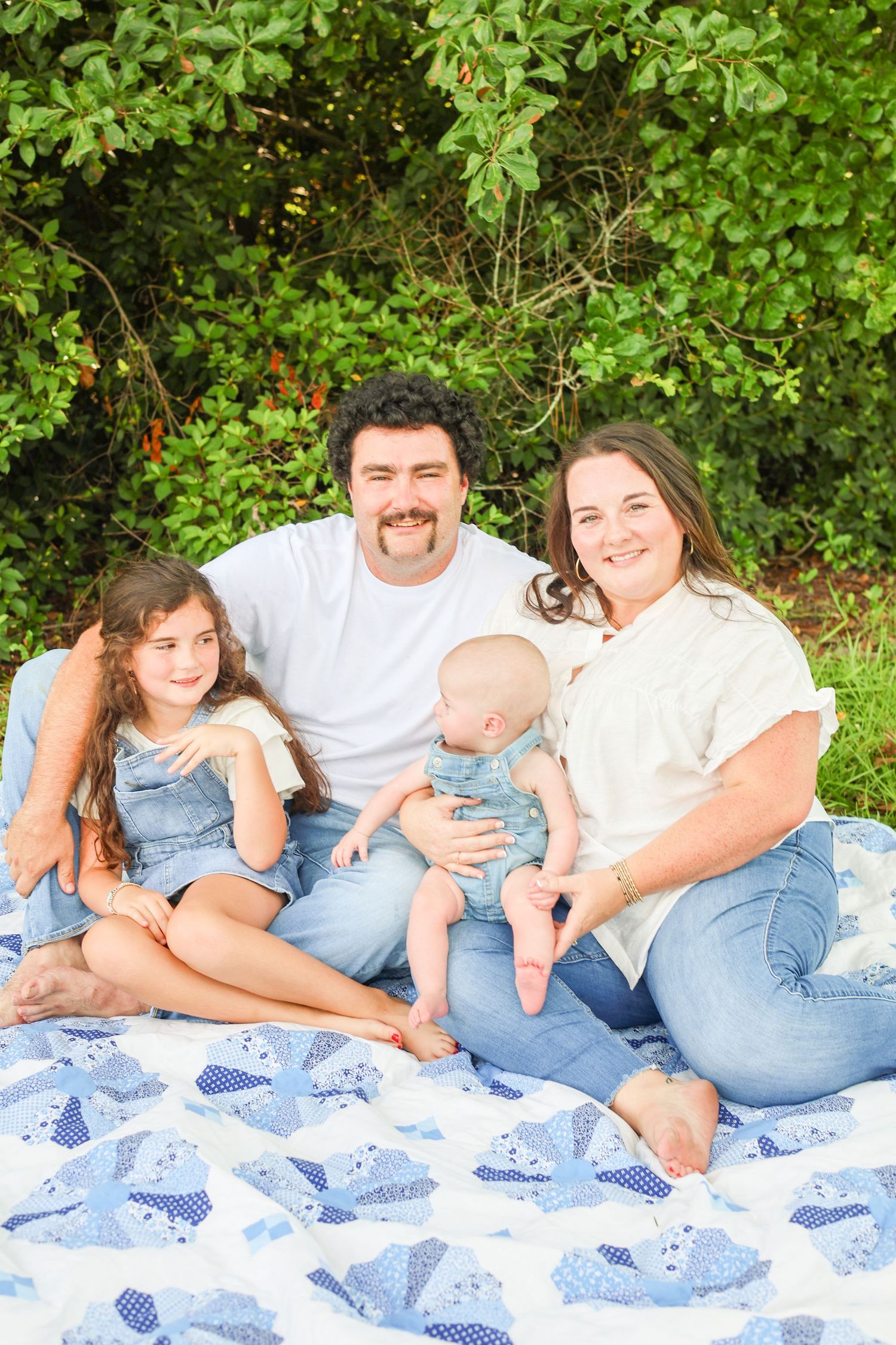 Happy family of four sitting on a blue quilt outdoors, parents with young daughter and baby, green trees background.