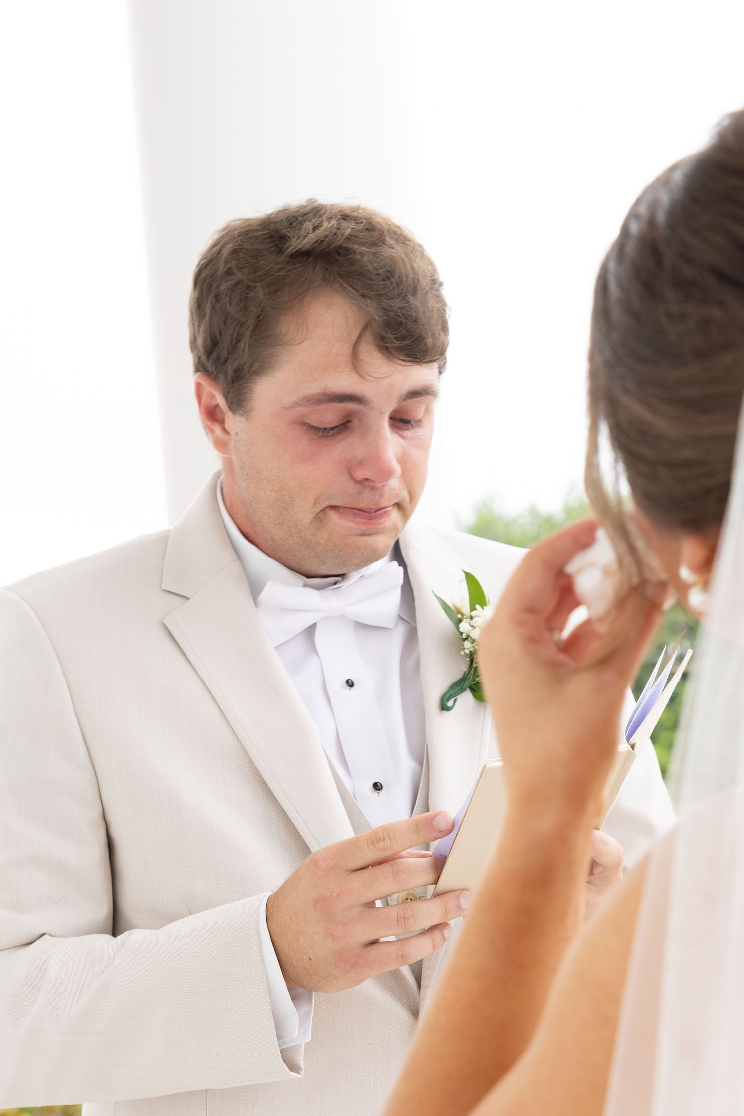 Emotional groom in white tuxedo crying while reading wedding vows during ceremony.
