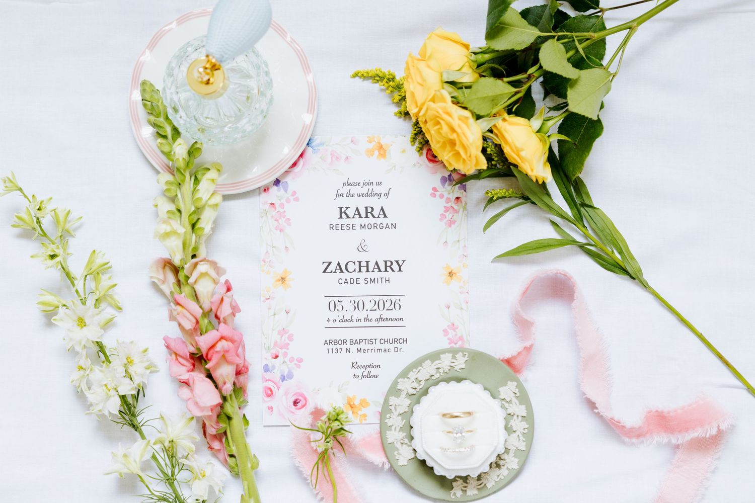 Elegant wedding invitation suite with floral details, yellow roses, pink ribbon, and decorative ring box on white linen.