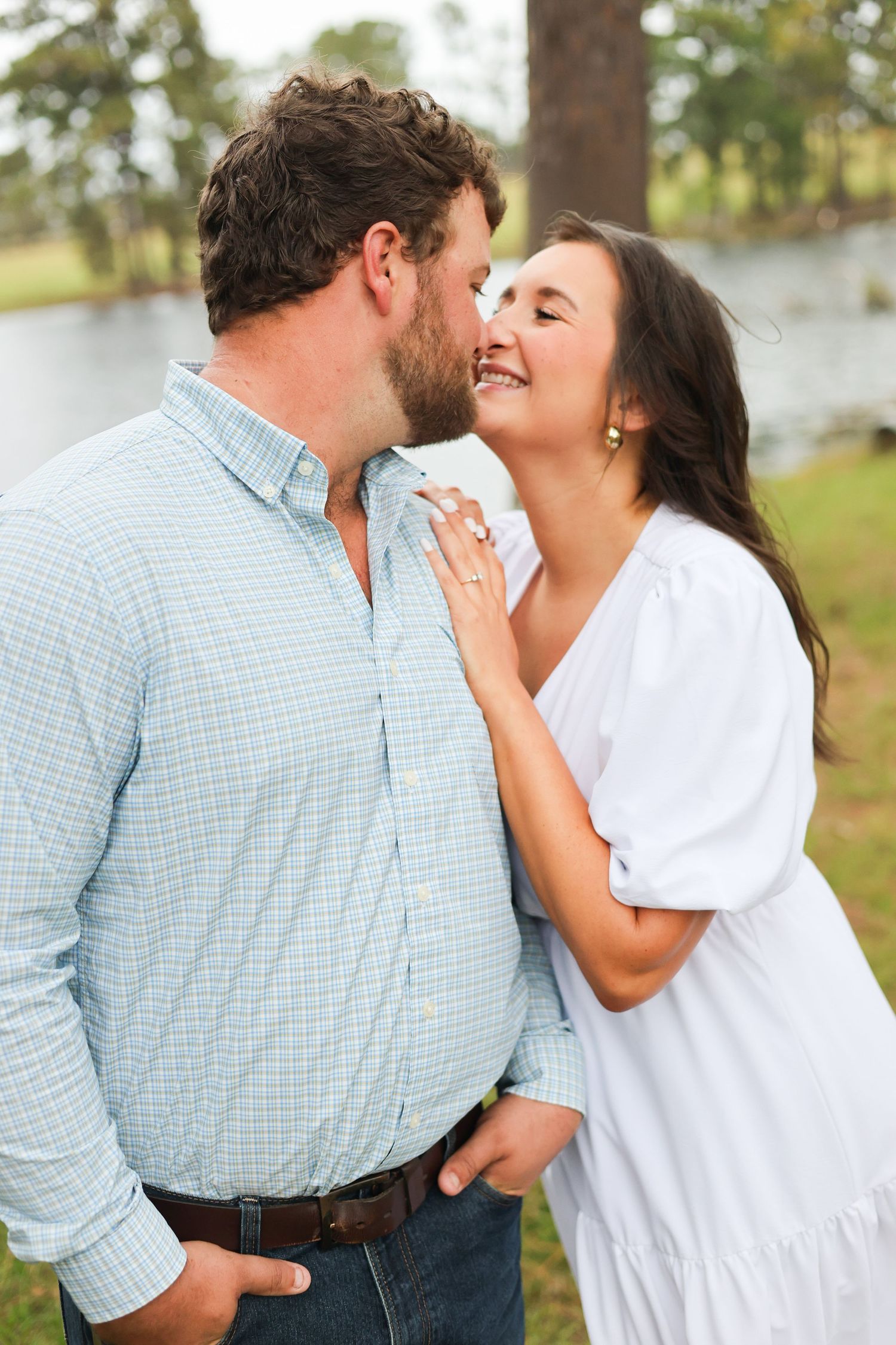 A couple sharing a tender moment outdoors in casual attire by a lake.
