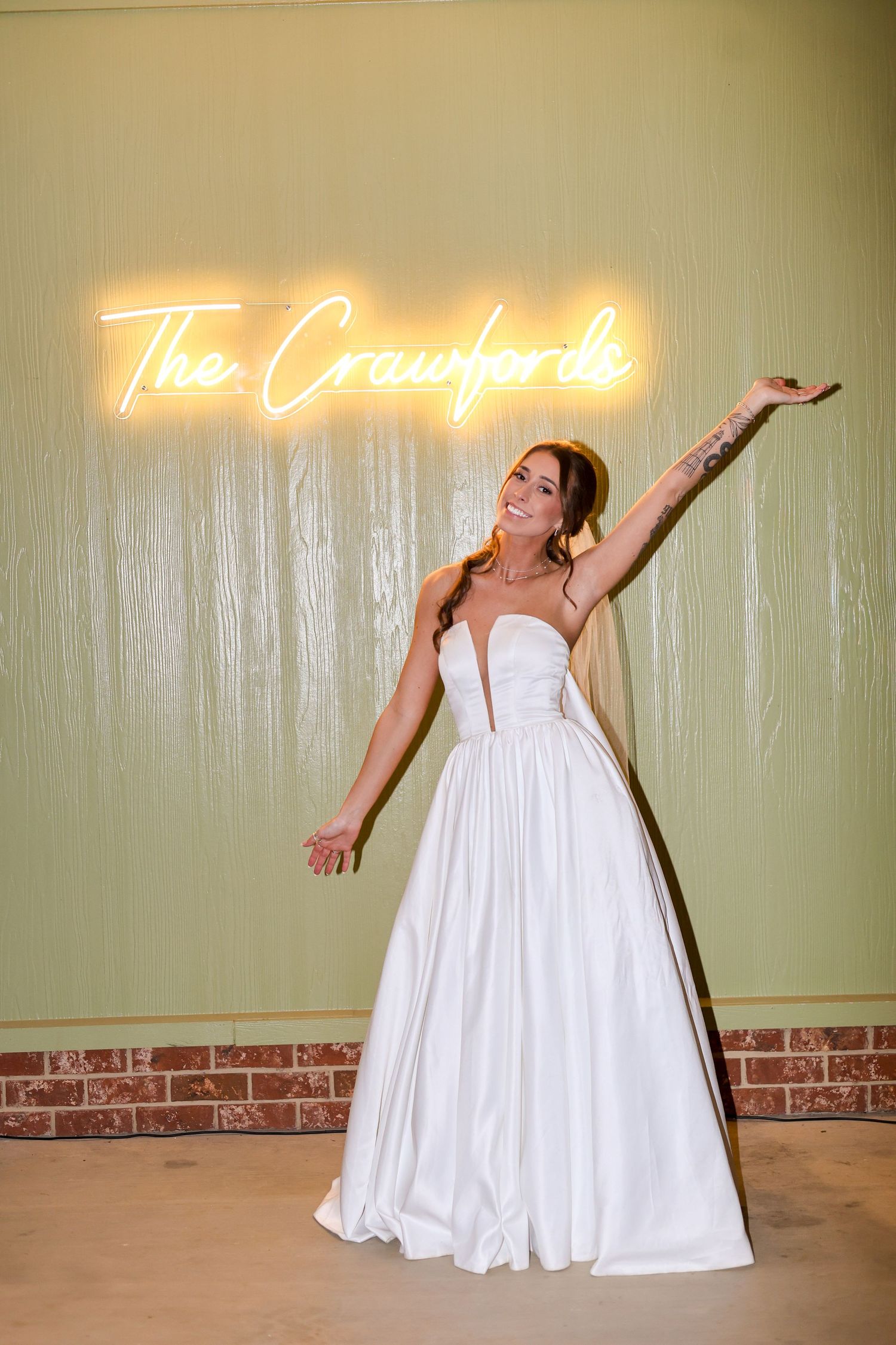 A bride in a white wedding gown poses joyfully under a neon sign that reads 'The Crawfords'.