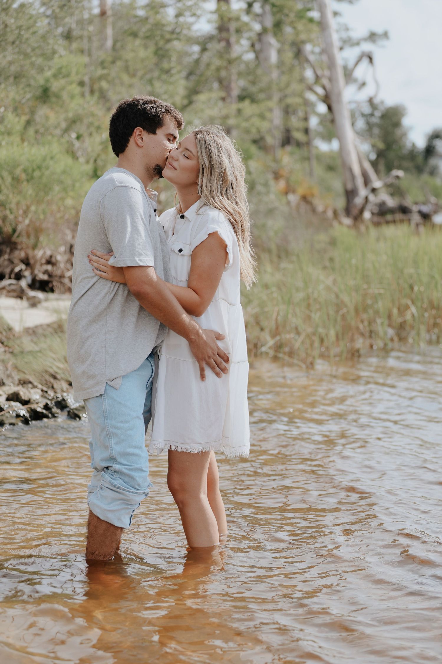 A romantic couple embraces while standing in shallow water near a wooded shoreline.