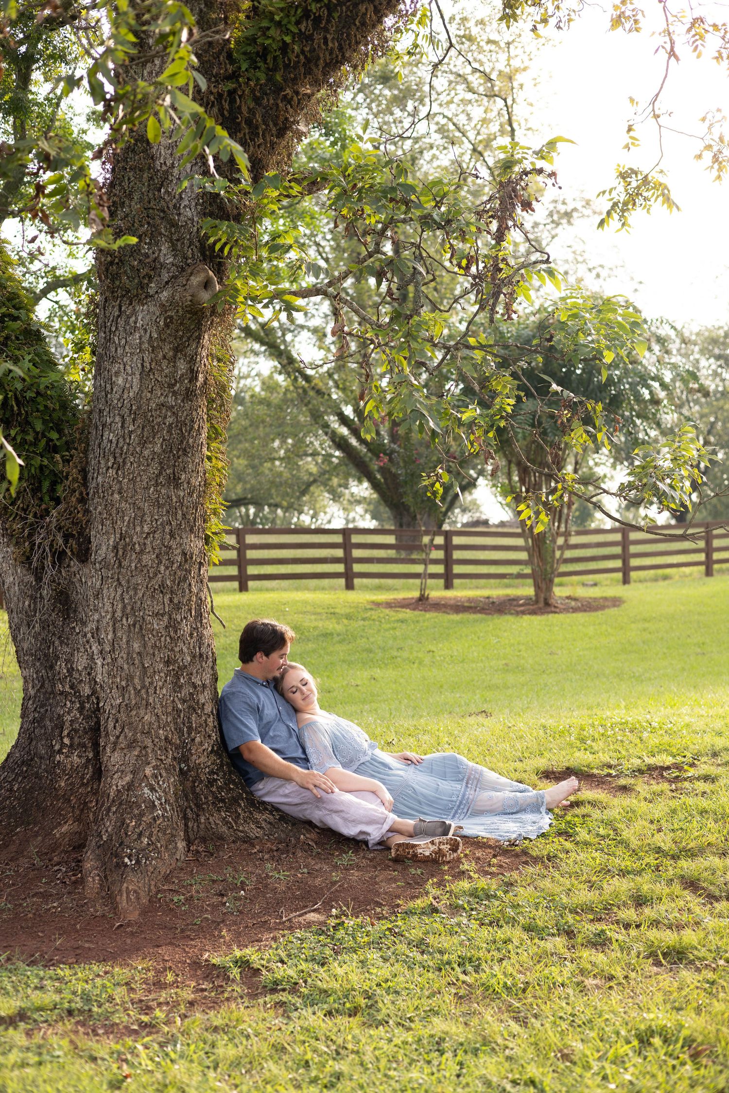 Couple relaxing under a large tree in a green park, leaning against the trunk on a sunny day.
