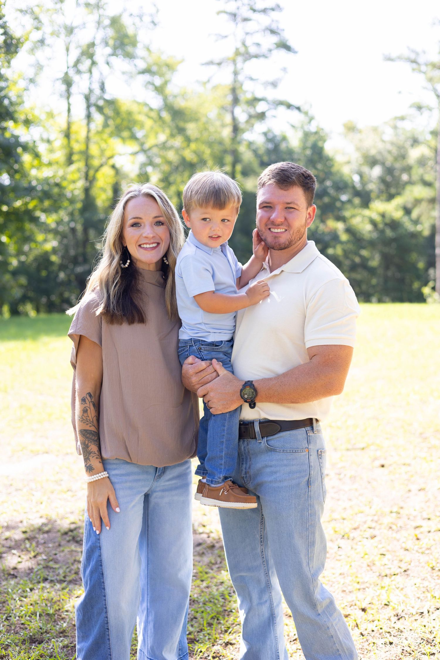 A happy young family with a toddler posing outdoors in a sunny green park, wearing casual jeans and neutral tops.