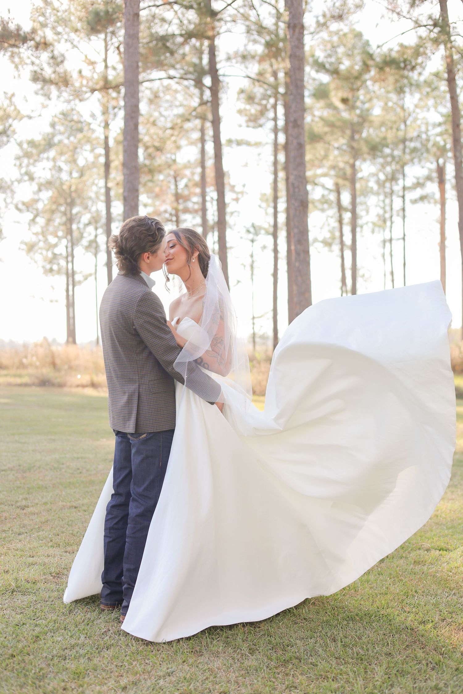 Bride and groom share a kiss outdoors with wedding dress flowing in the wind, surrounded by tall pine trees.