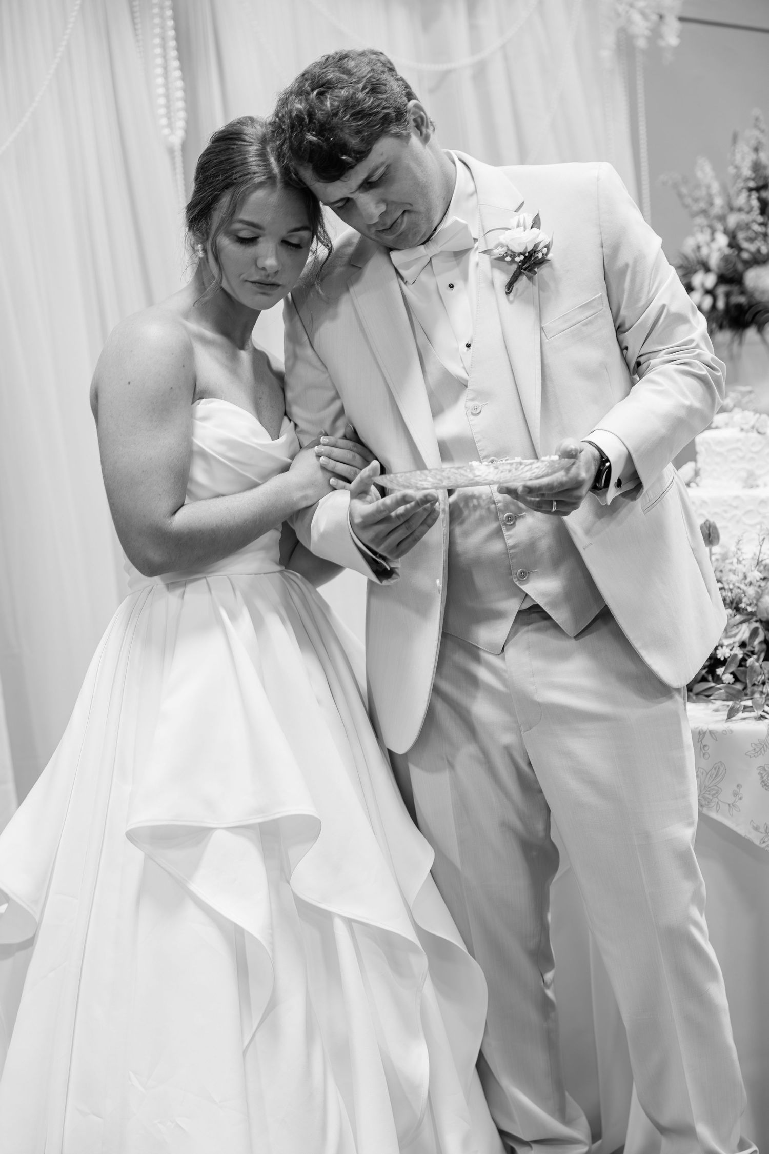 Black and white photo of bride and groom in elegant attire reading during wedding reception cake cutting.