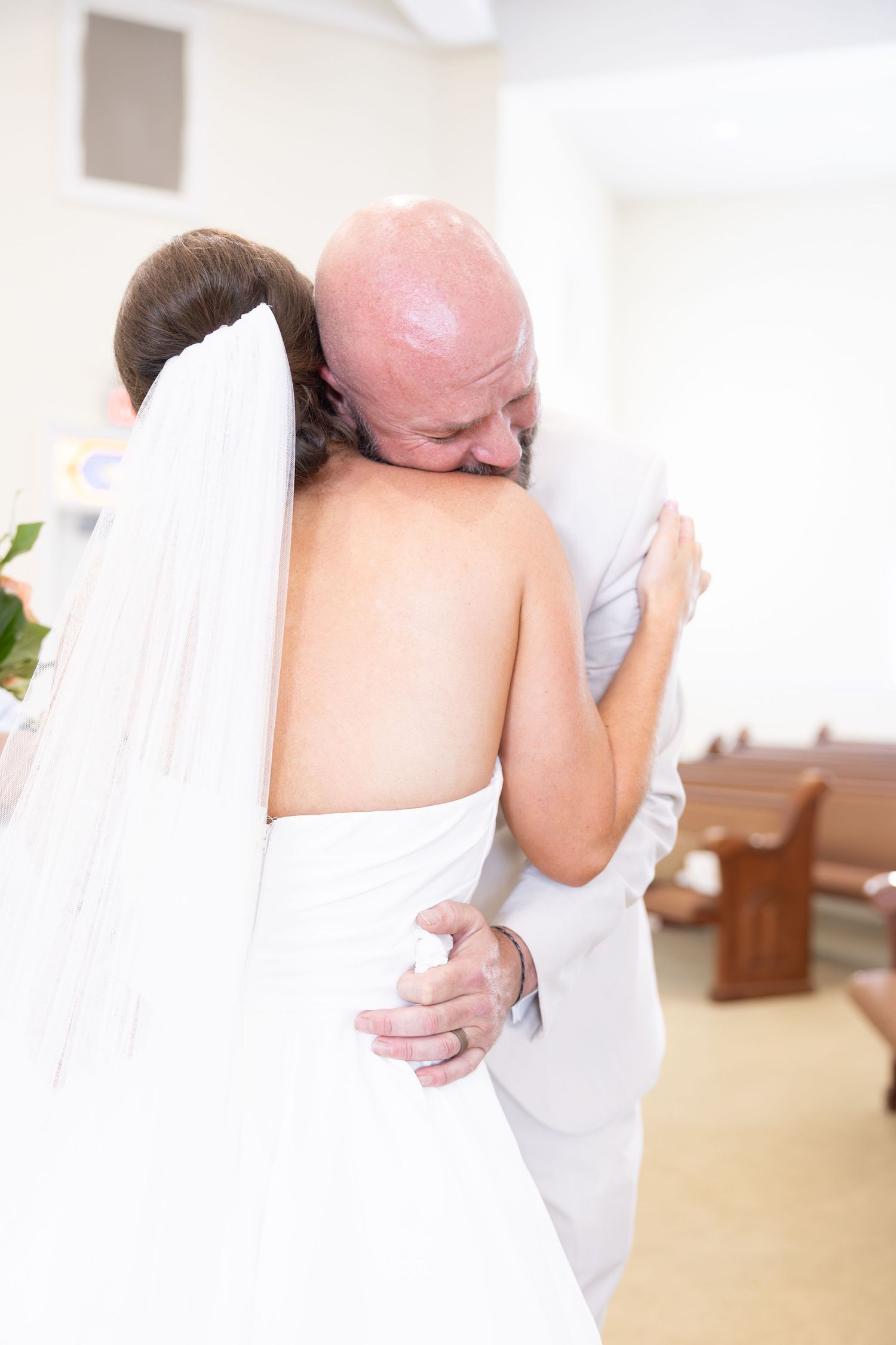 Emotional father of the bride hugging his daughter in her white wedding gown and veil inside a church.