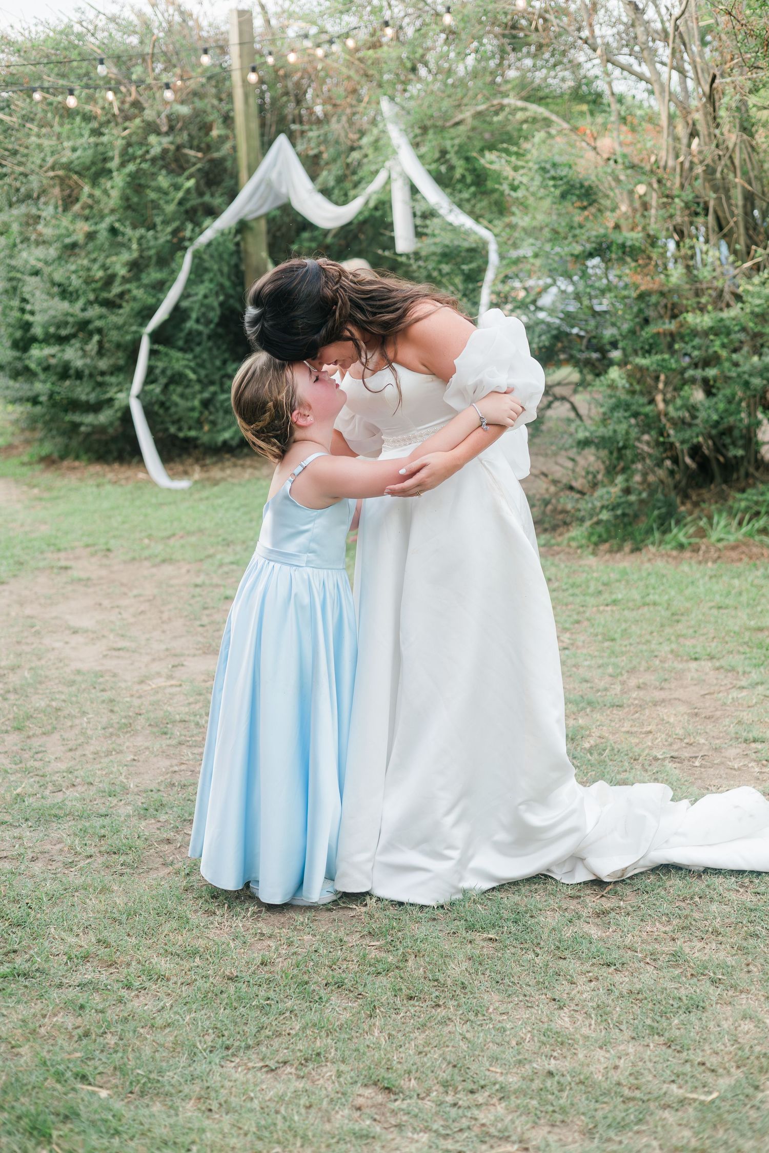 Bride in white gown sharing tender moment with flower girl in light blue dress at outdoor wedding.