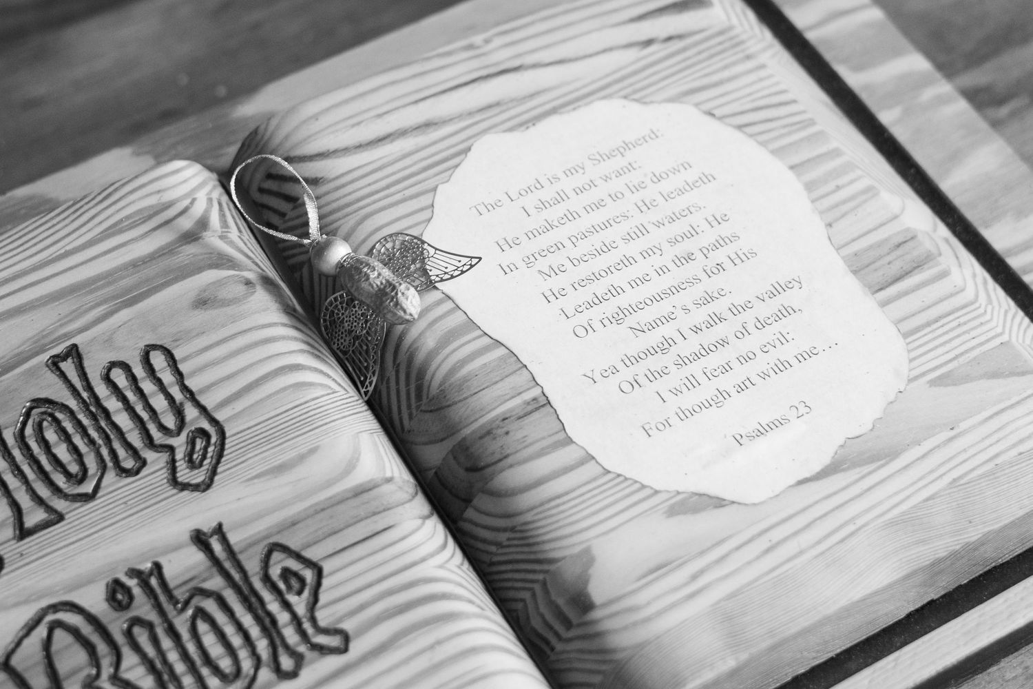 Black and white photo of a Holy Bible with a Psalm 23 card and decorative cross on a wooden surface.
