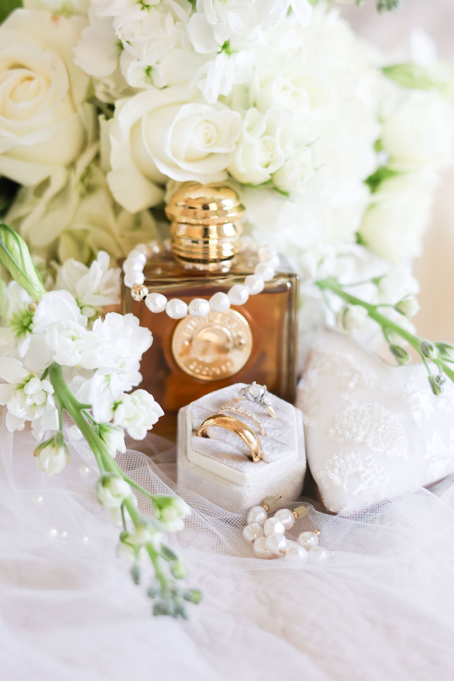 Elegant wedding details with white roses, delicate pearls, and golden honey jars arranged on white lace fabric.