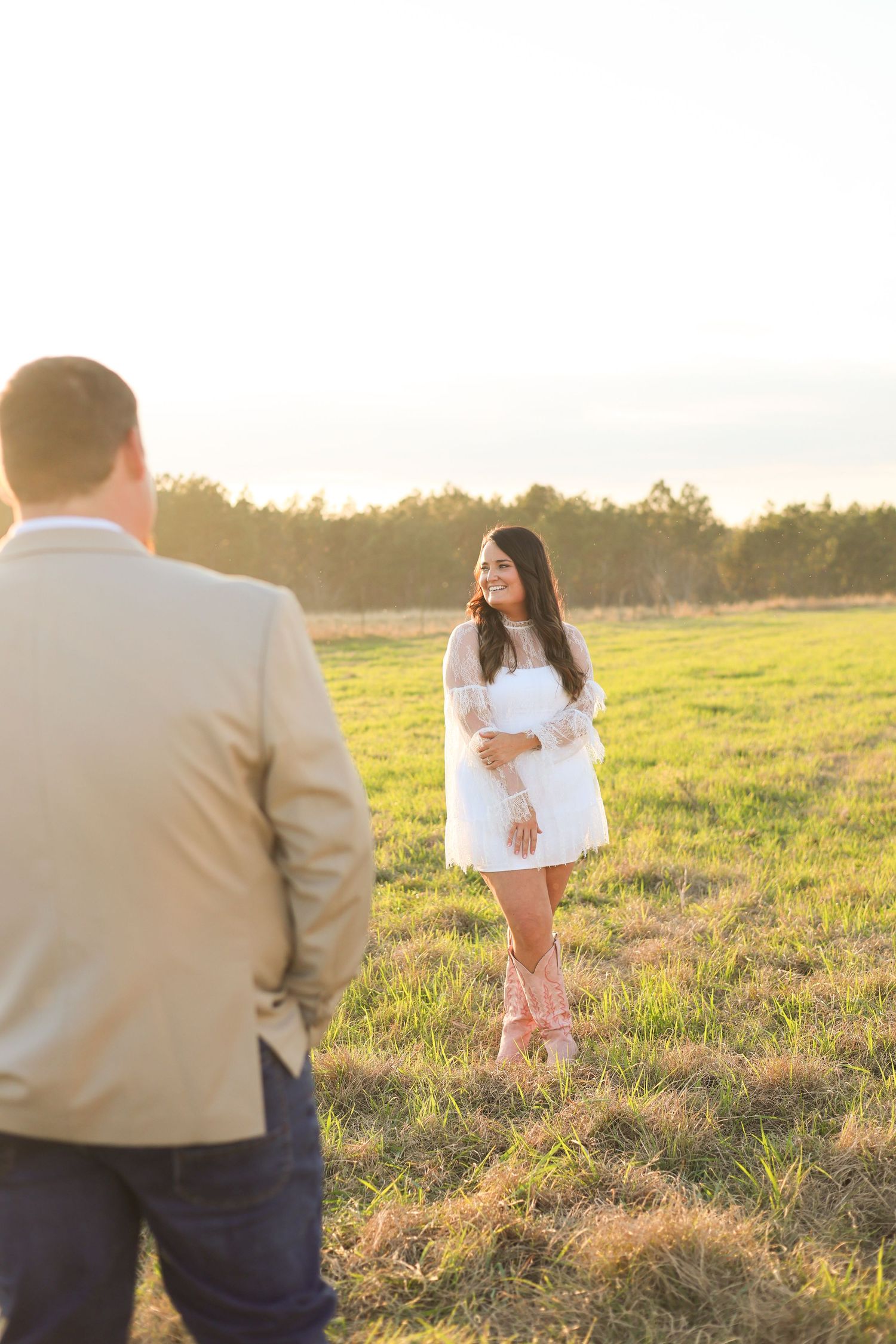 A couple shares a romantic moment in a sunlit field at golden hour, with her wearing a white dress and pink boots.