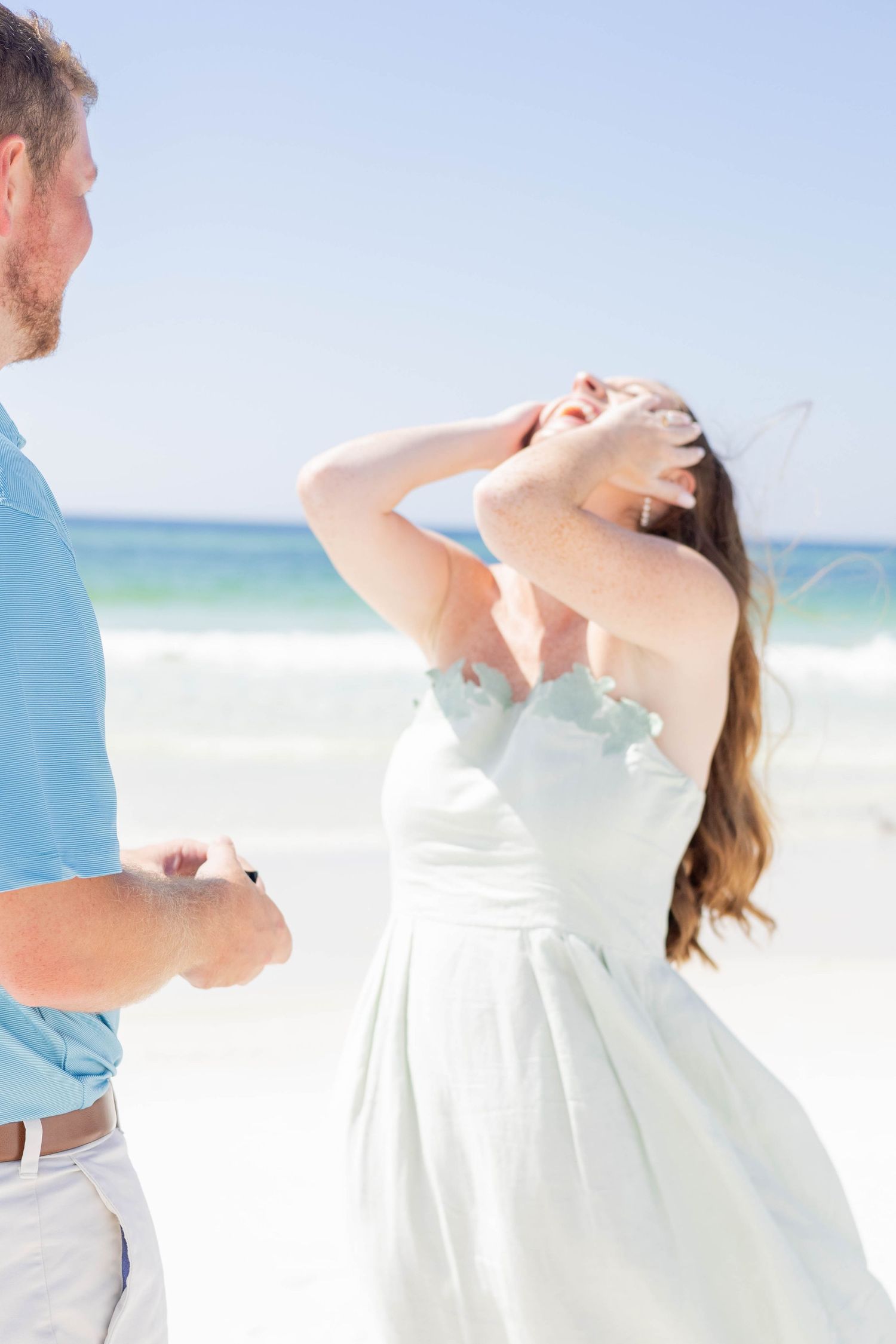 Couple sharing a romantic beach proposal moment, woman in white dress with hands on face, ocean backdrop.