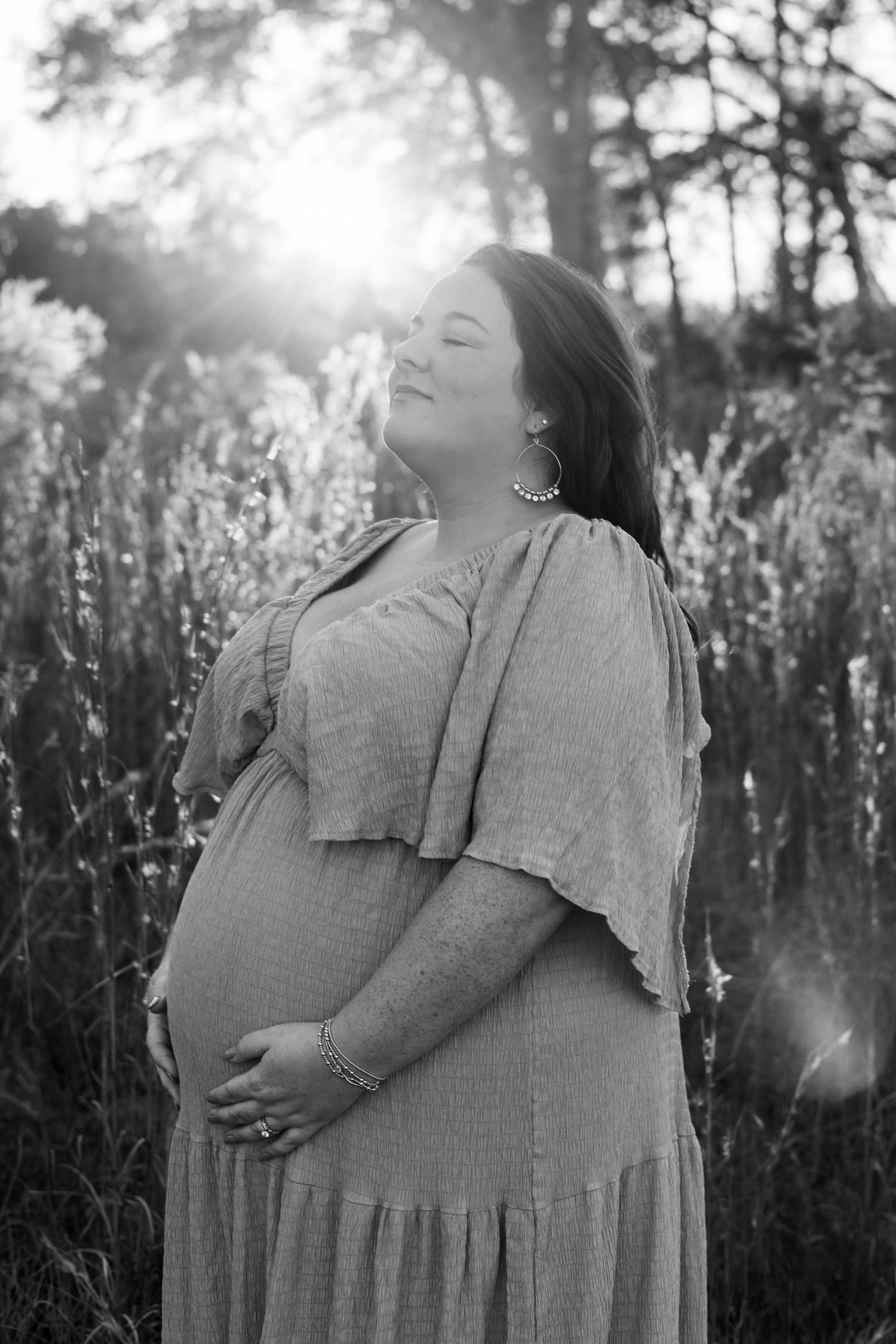 Black and white maternity photo of a pregnant woman in a flowy dress, cradling her belly outdoors with sunlight glowing behind her.