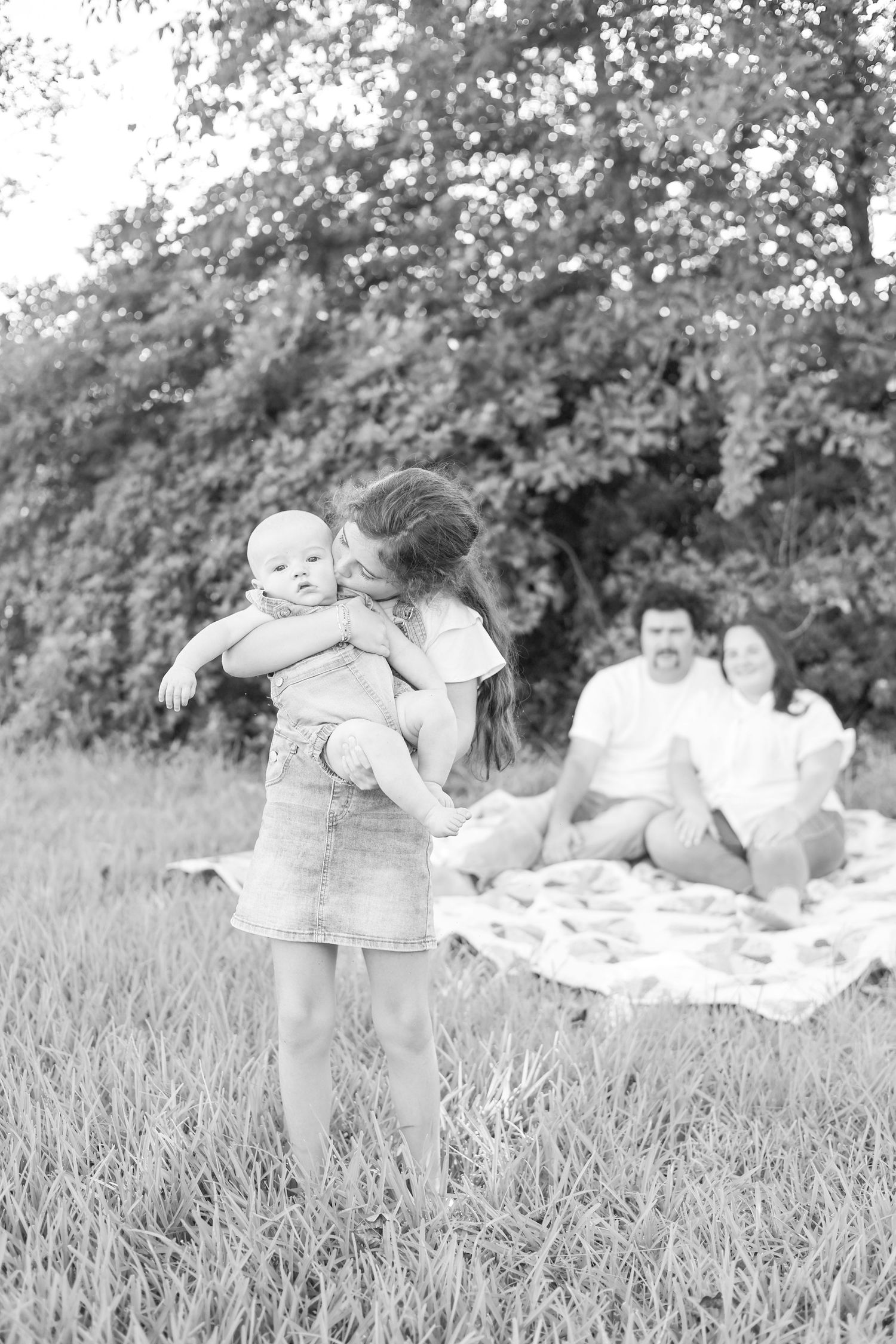 Black and white photo of a girl holding a baby outdoors while parents relax on a blanket in a grassy field.