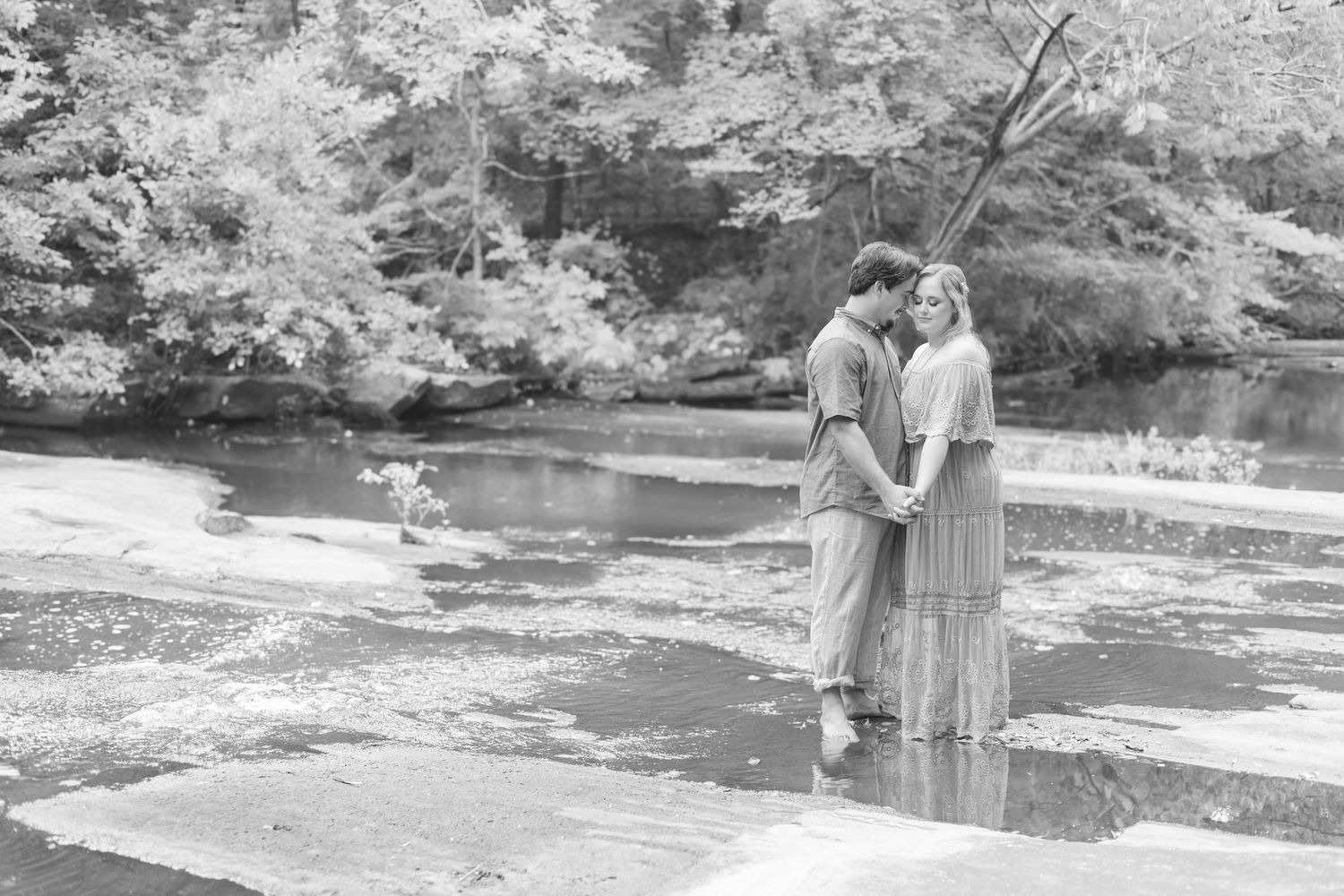 Black and white photo of a couple holding hands near a serene creek surrounded by lush trees.