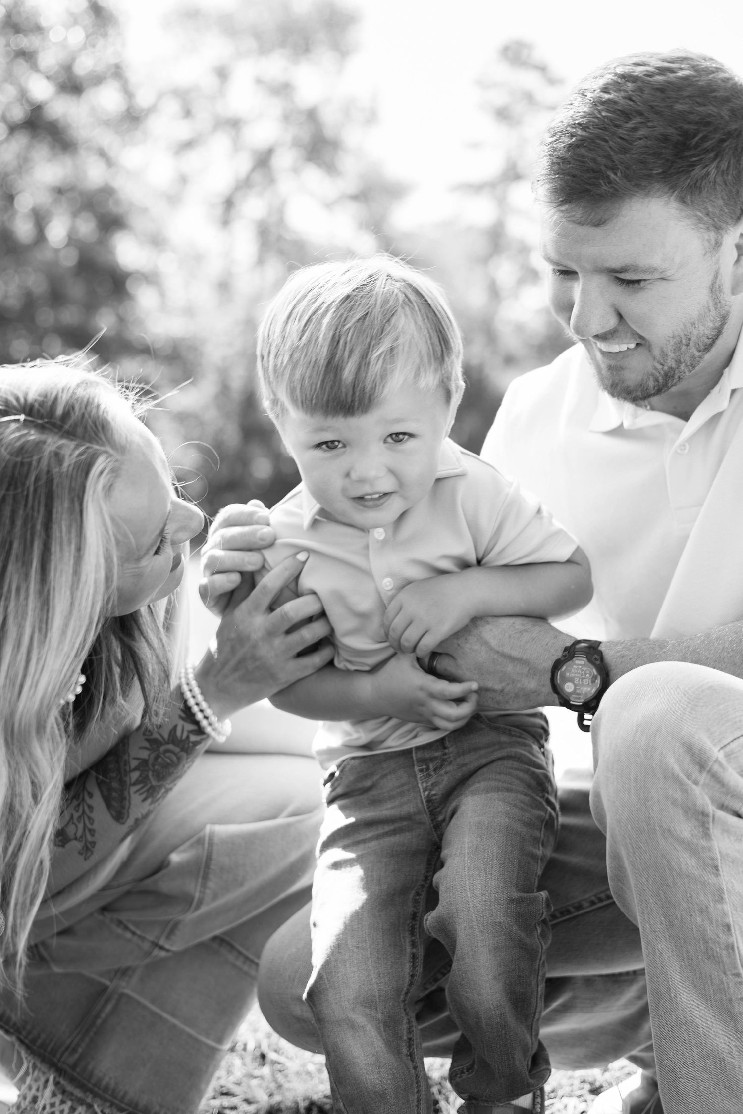 Black and white photo of a happy family with parents smiling at their toddler son outdoors in natural light.