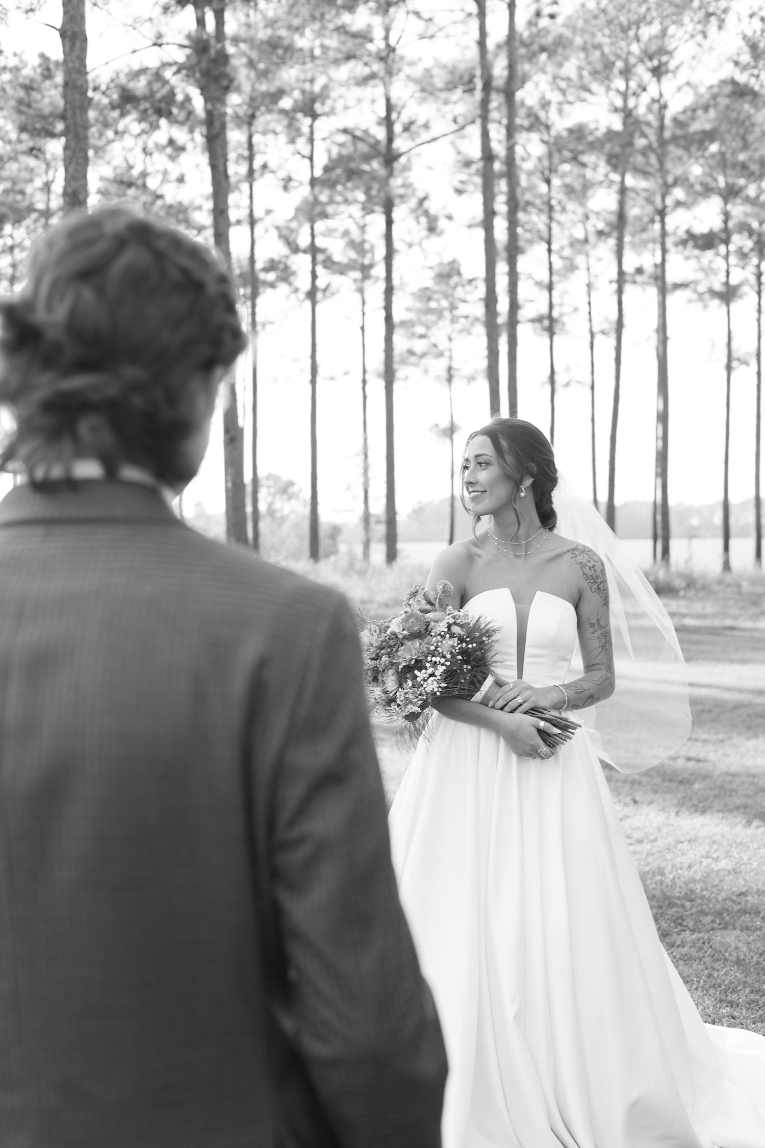 Black and white photo of a smiling tattooed bride in a white gown holding a bouquet during an outdoor first look.