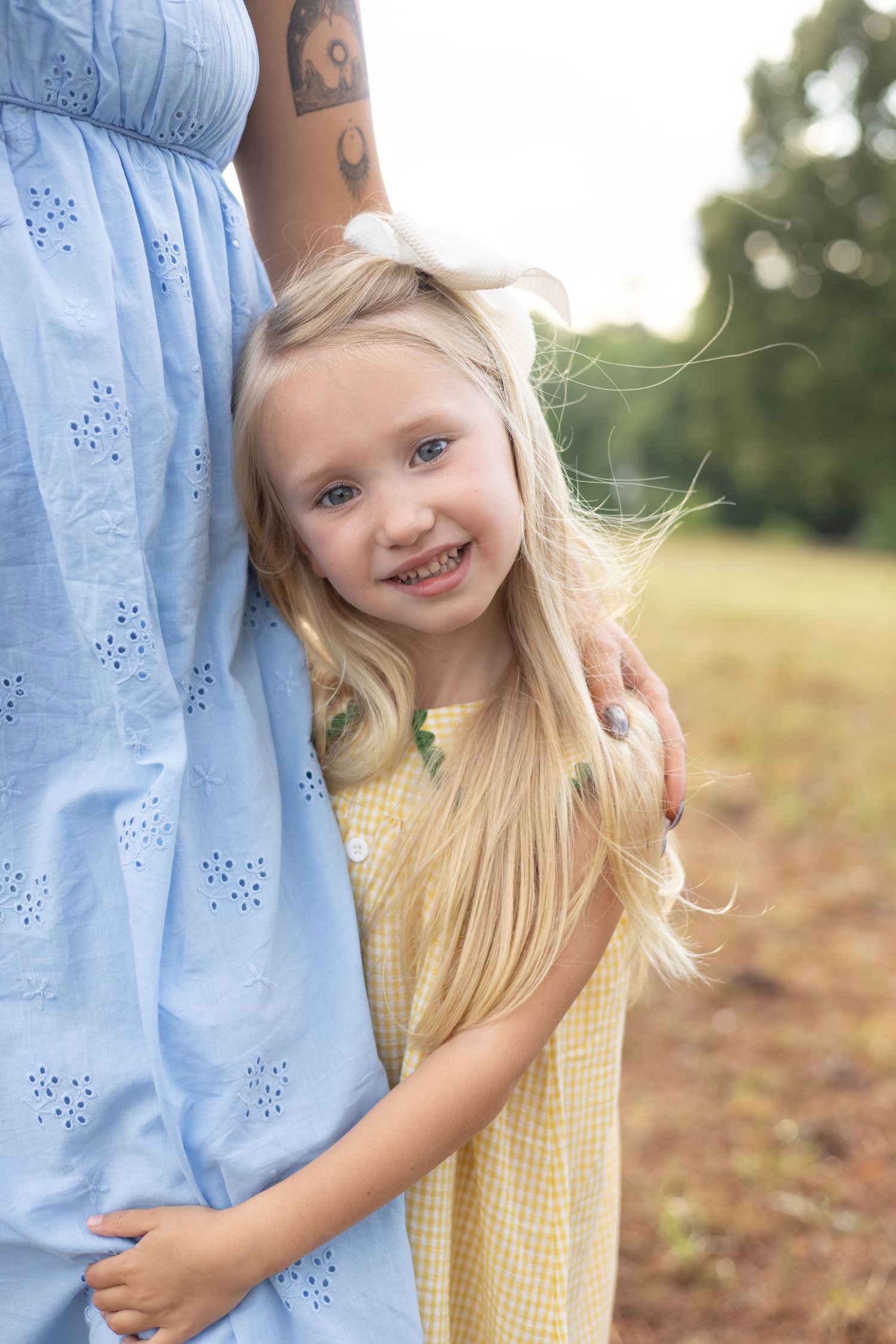 A young blonde girl in a yellow dress smiles while hugging her mother in a blue dress outdoors.