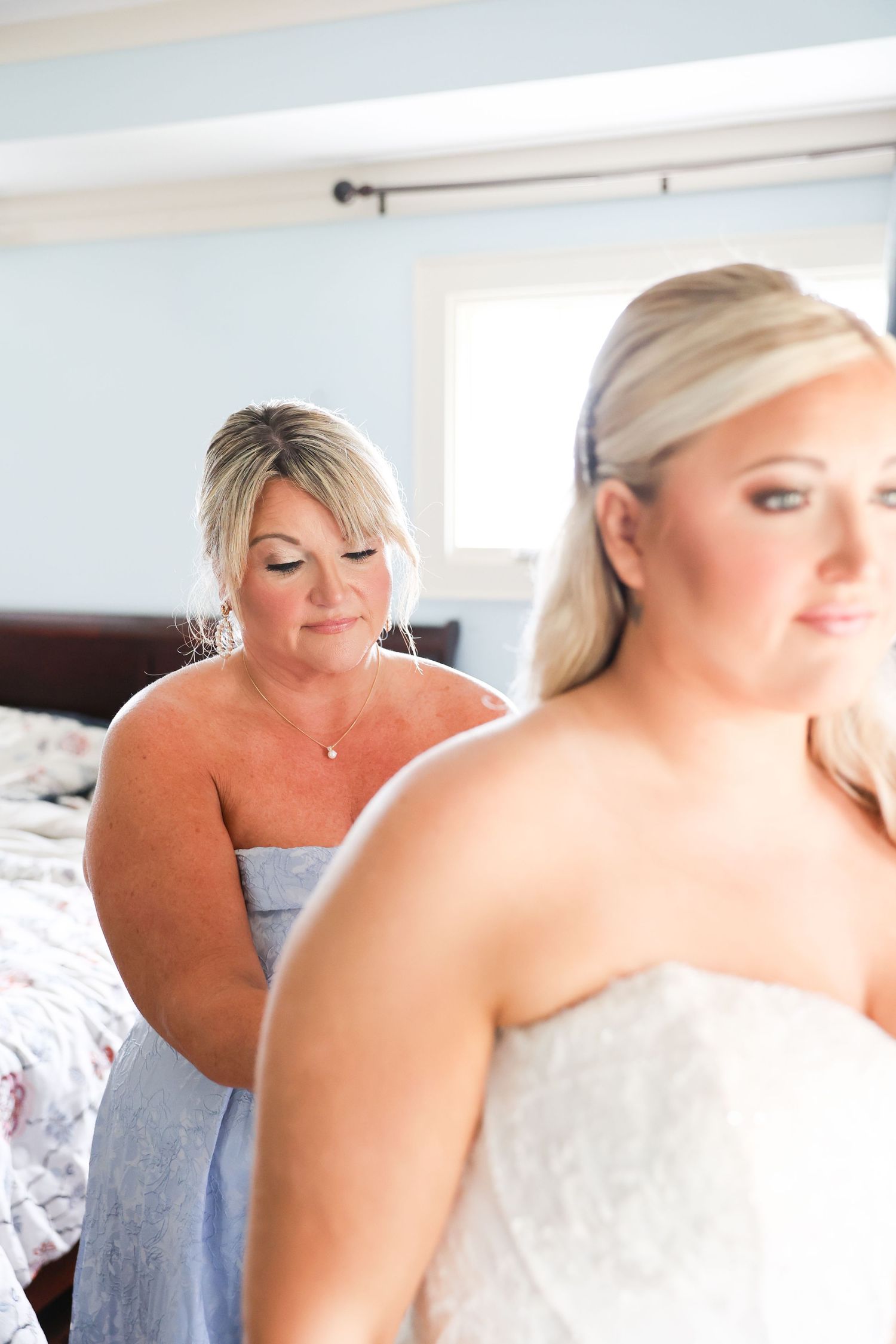 A mother helps her daughter button up a white lace wedding gown in a softly lit bedroom.