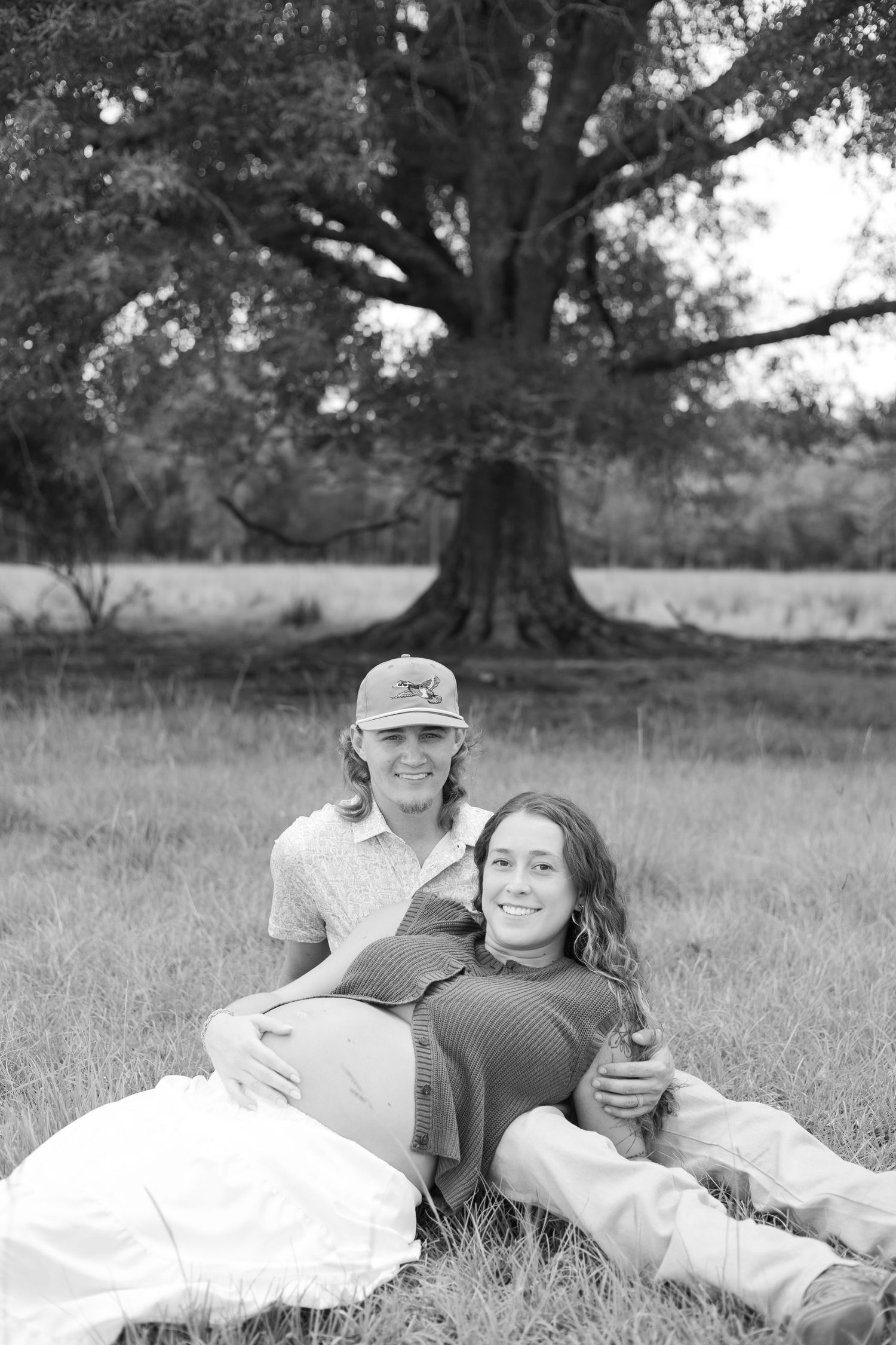Black and white maternity photo of couple sitting in grass field with large oak tree backdrop.