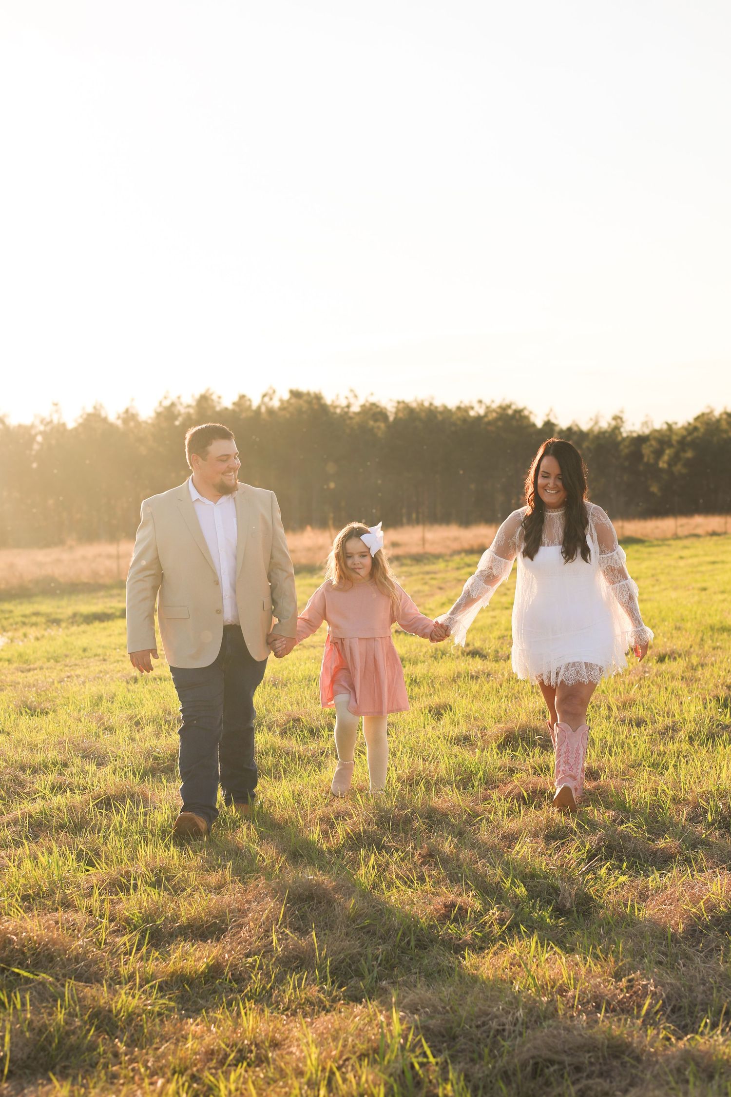 A family walks together holding hands in a sunlit meadow during golden hour as sunlight streams through trees.
