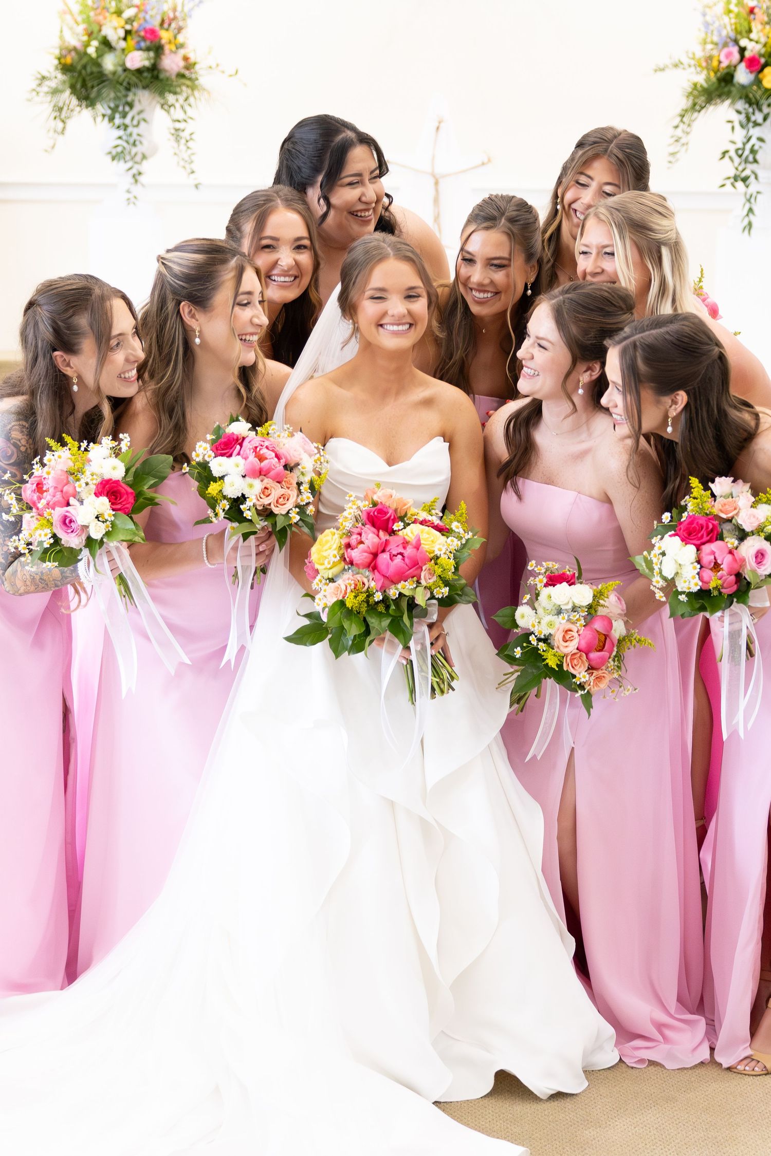 Smiling bride in white gown surrounded by bridesmaids in pink dresses holding colorful bouquets.