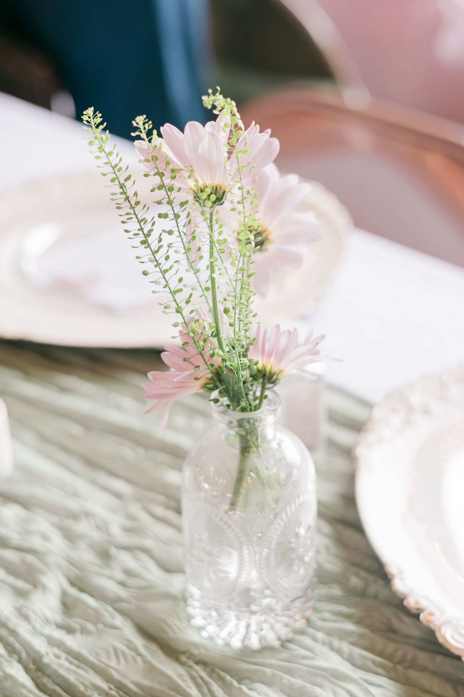 Small glass vase with pink daisies and greenery as elegant wedding table centerpiece decoration.