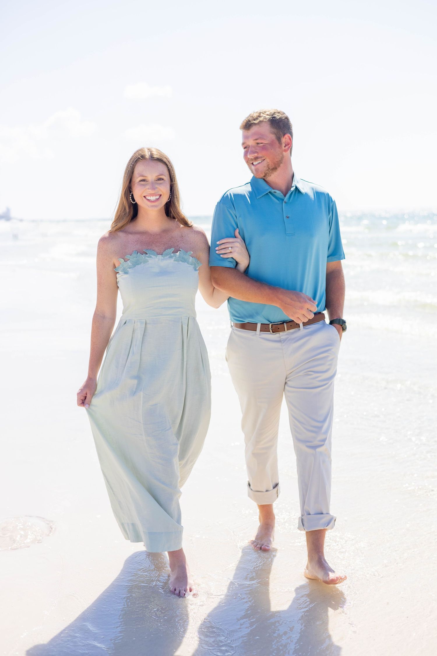 A couple walks barefoot together along a sunny beach, wearing light summer attire.