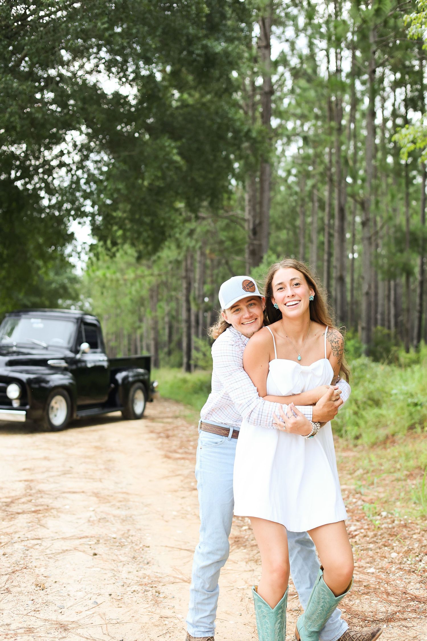 A playful couple embraces on a dirt road surrounded by tall pine trees and a black truck in the background.