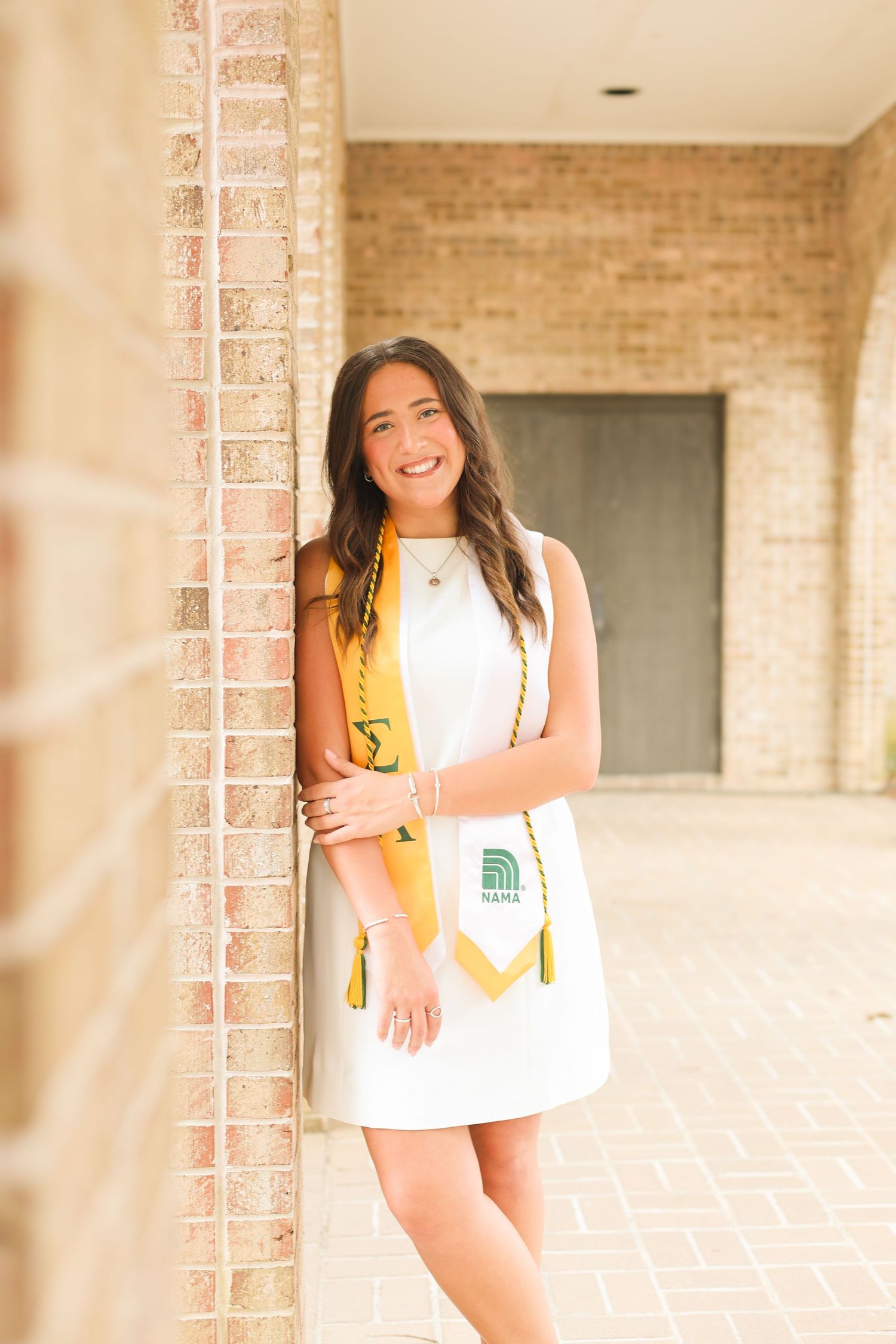 Indoor graduation photos featuring white dress and gold stole against brick wall backdrop.