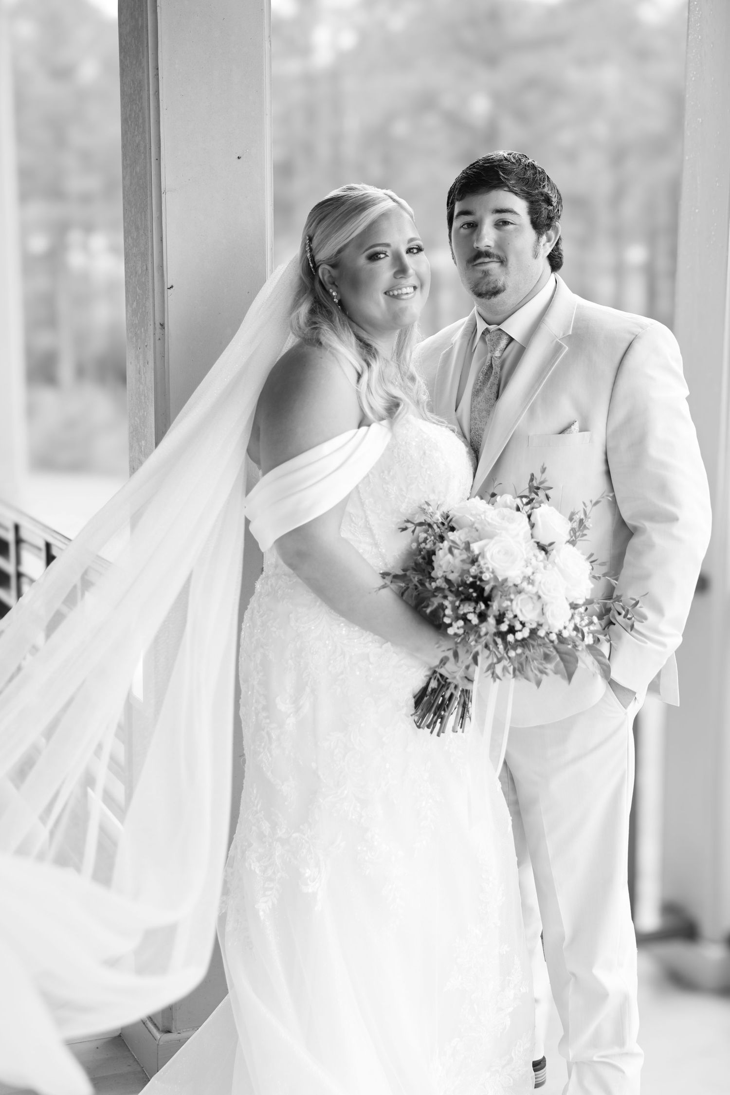 Black and white wedding portrait of bride holding bouquet and groom in light suit posing together indoors.