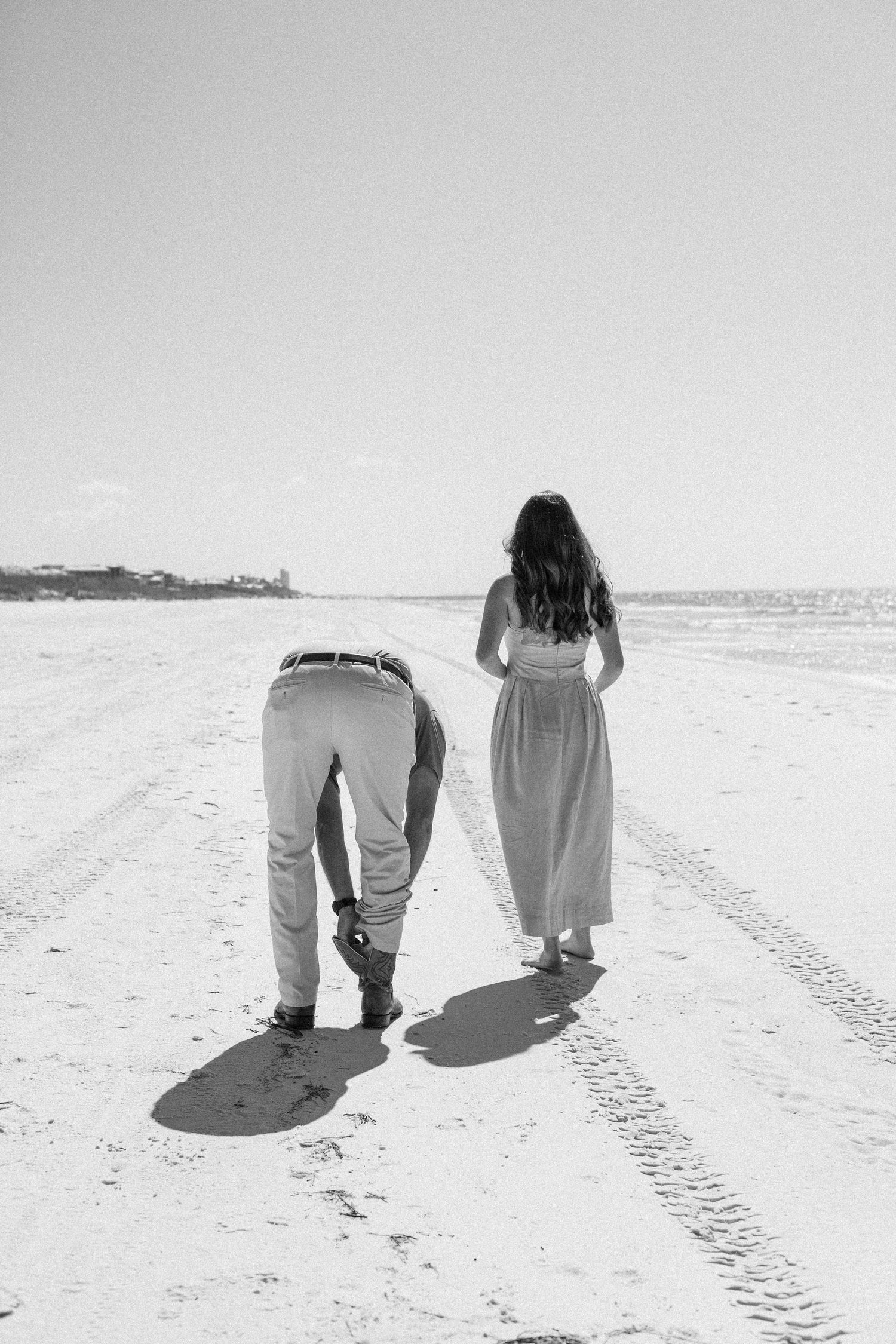 Black and white photo of a couple walking on a sandy beach, woman in long dress, man bending down.