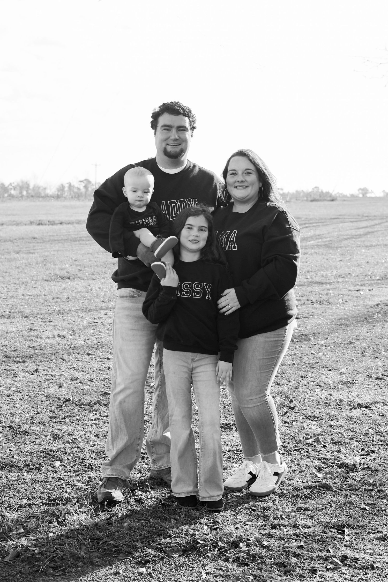 Black and white family portrait of parents with young daughter and baby boy posing in an open field.