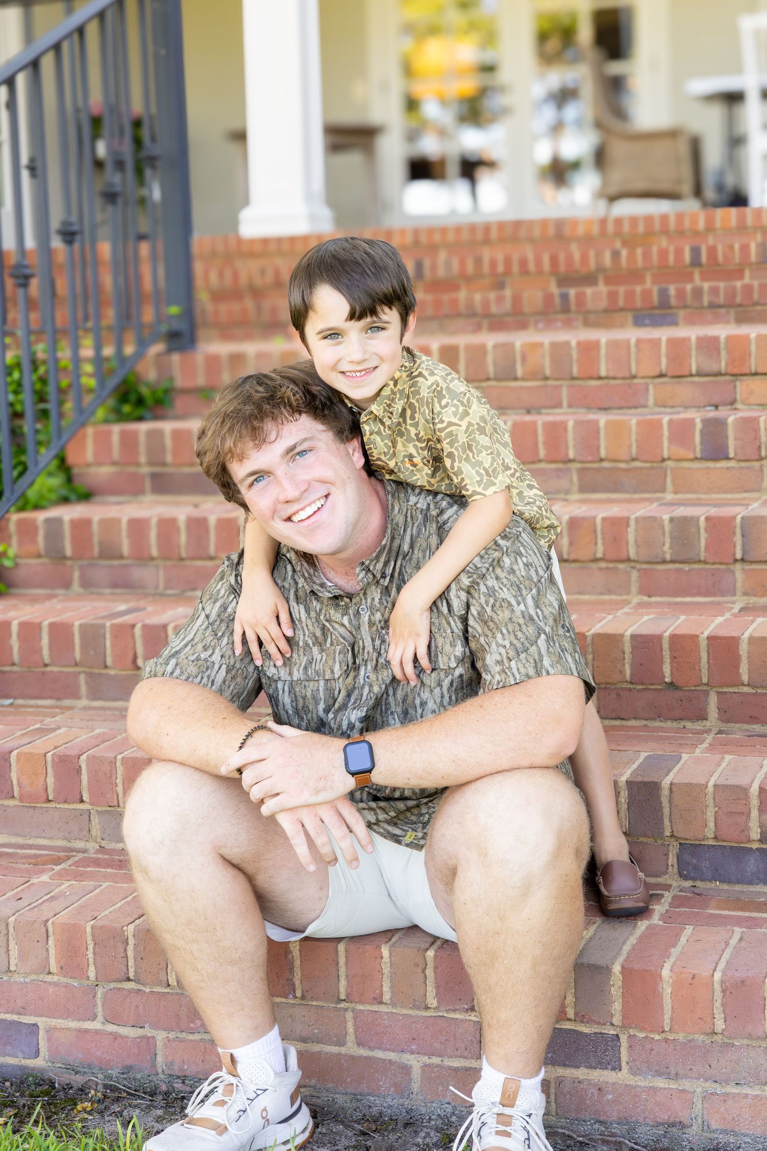 Father and young son smiling together on brick front porch steps of a house on a sunny day.