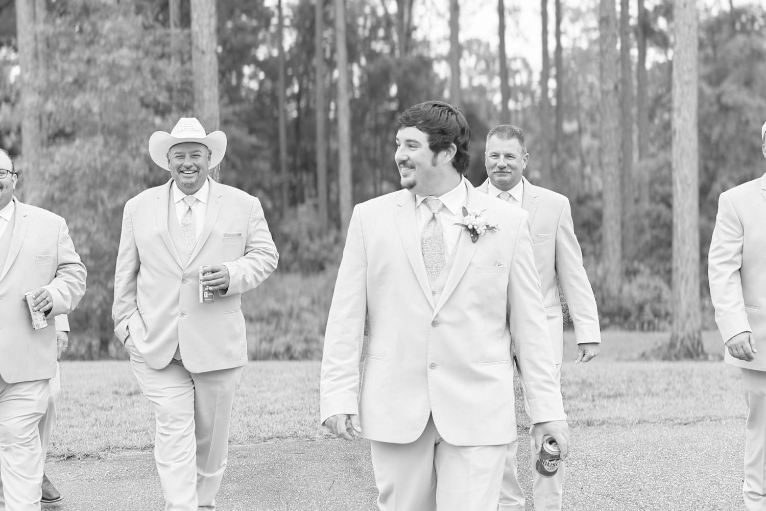 Groomsmen in light suits walk outdoors, smiling, with one wearing a cowboy hat, at a rustic wedding celebration.