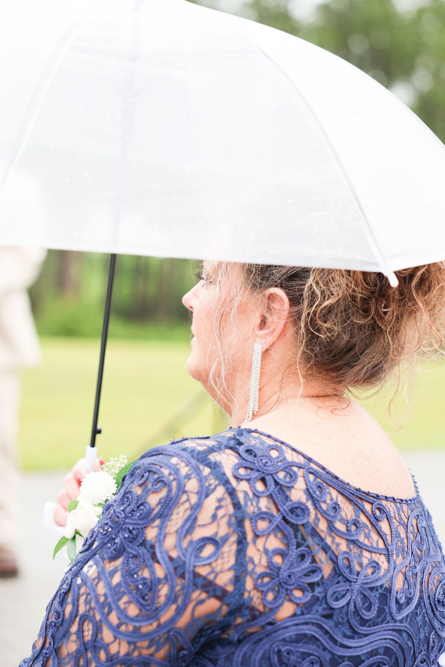 Woman in navy blue lace dress holding clear umbrella in rain at outdoor wedding ceremony.