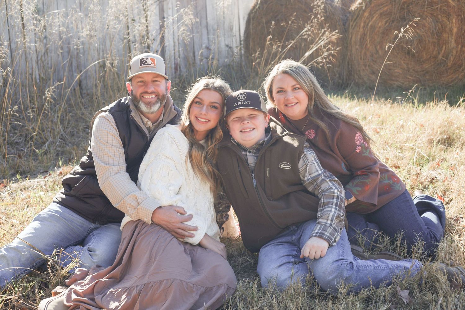 A family group sitting together outdoors in casual attire with round hay bales visible in the background.