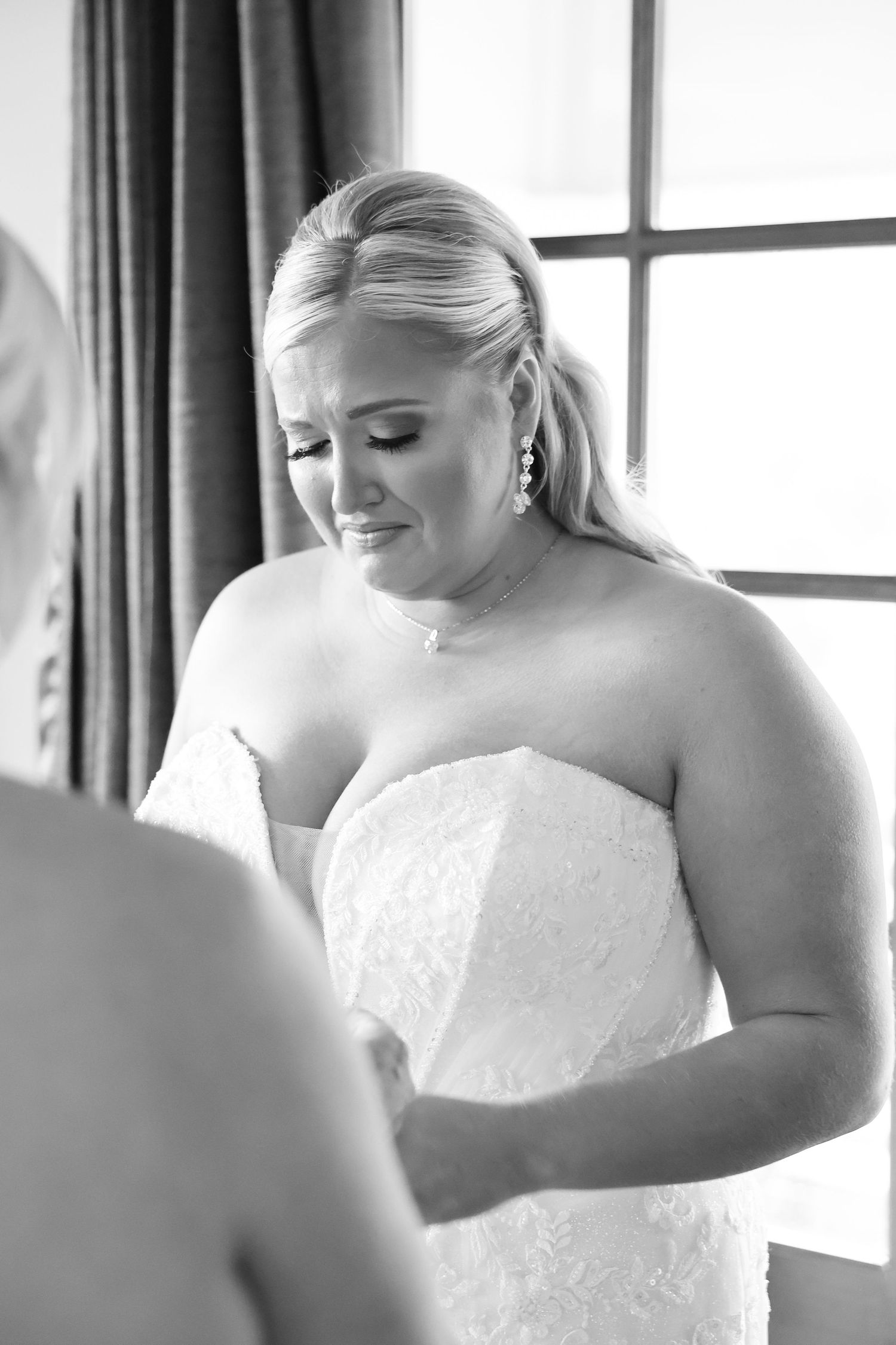 Emotional bride in strapless lace wedding gown crying while getting dressed in black and white photo.