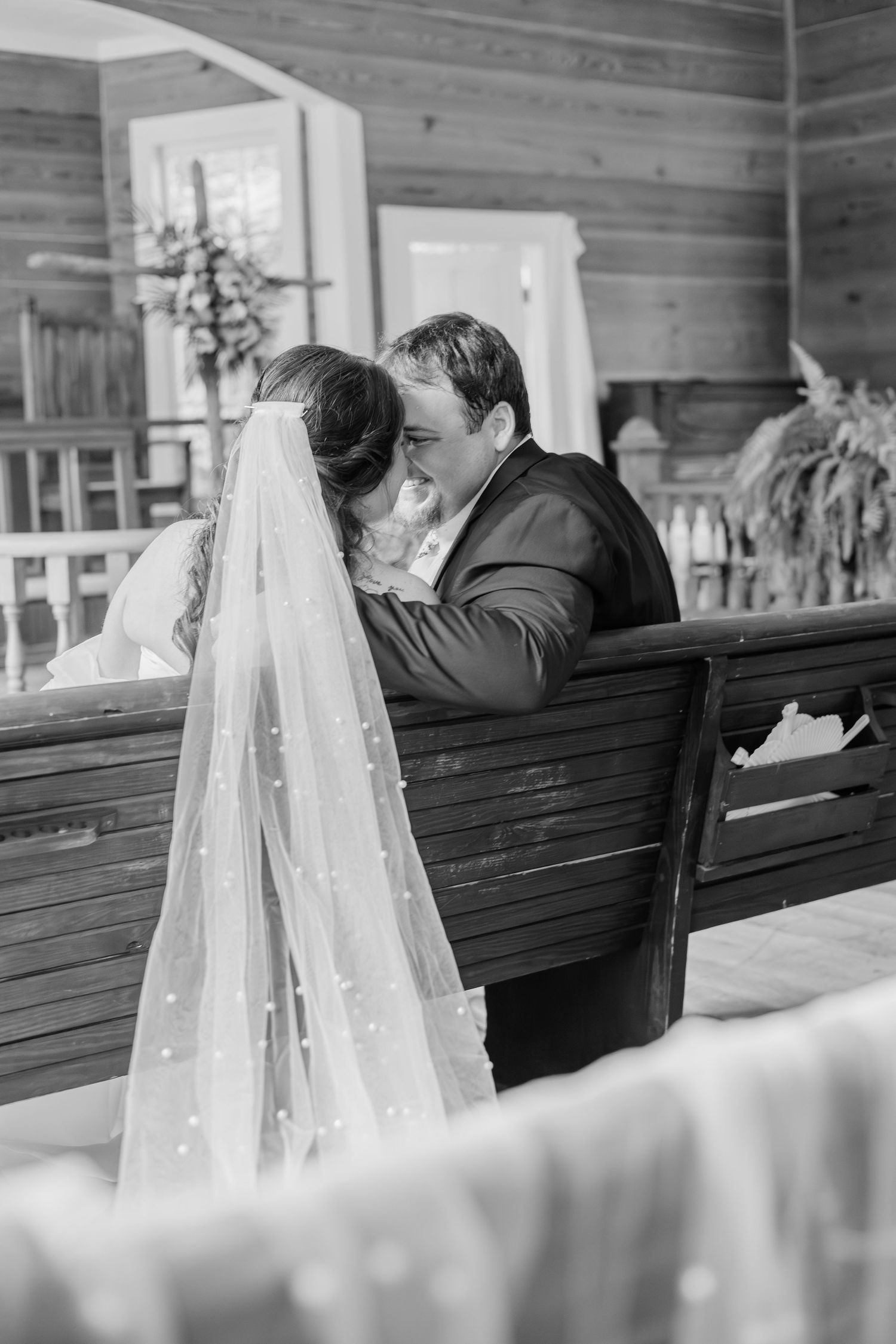 Bride and groom sharing intimate moment on wooden pew with pearl-adorned veil in black and white.