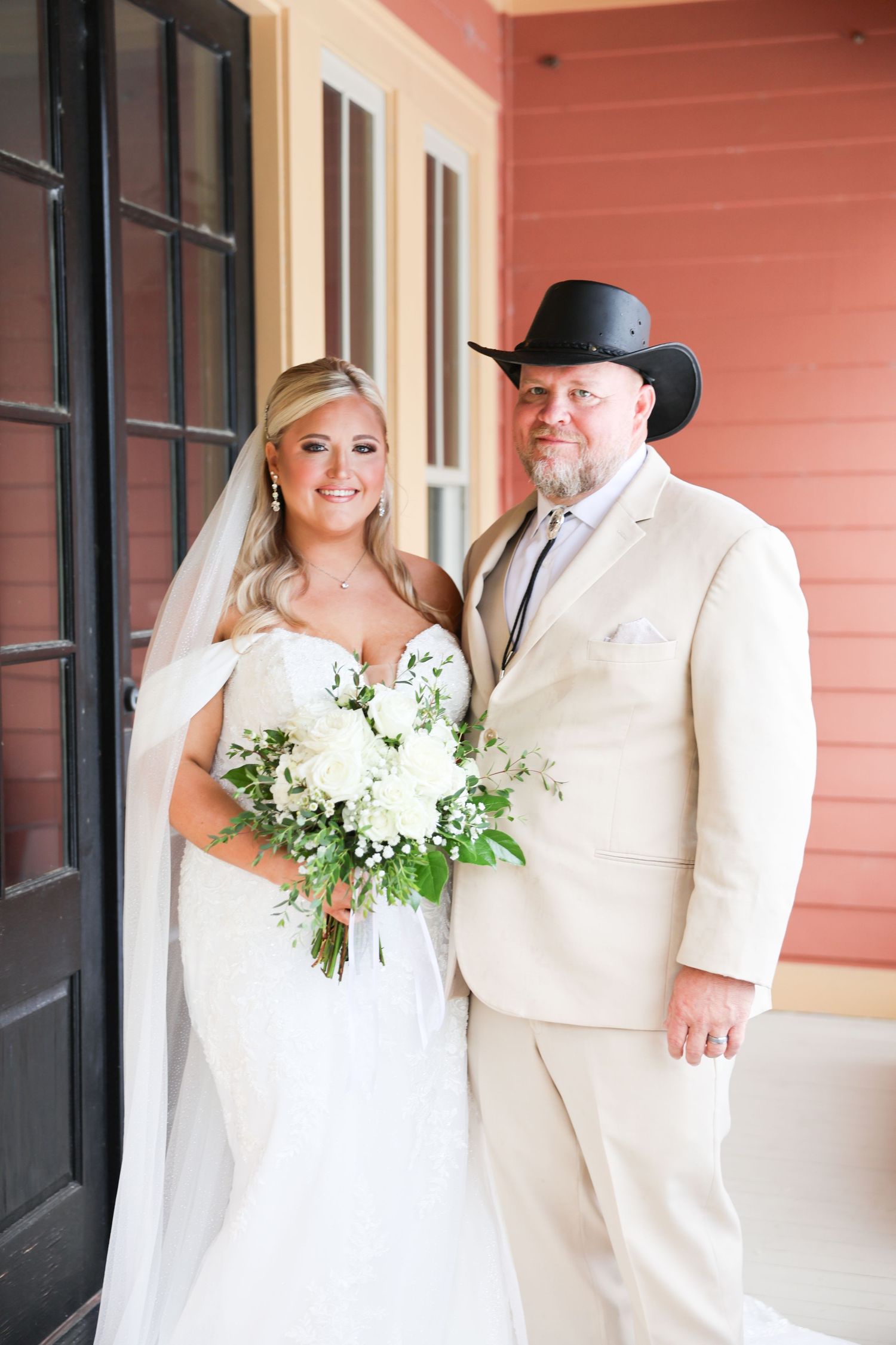 Bride in white lace gown with bouquet posing with father in tan suit and black cowboy hat.