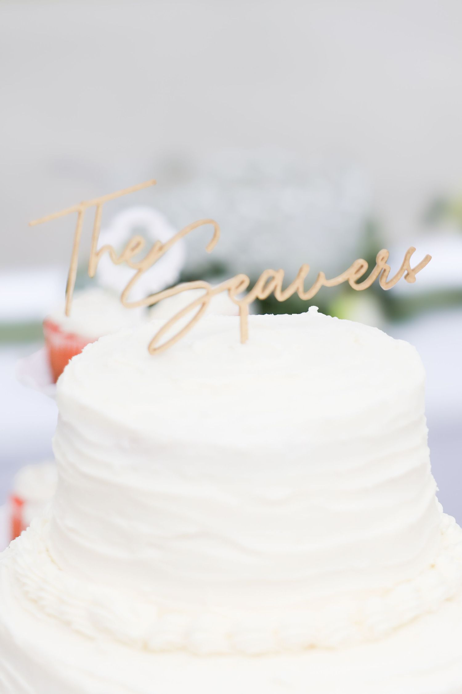 White wedding cake with gold laser cut 'The Seavers' cake topper in elegant script font.