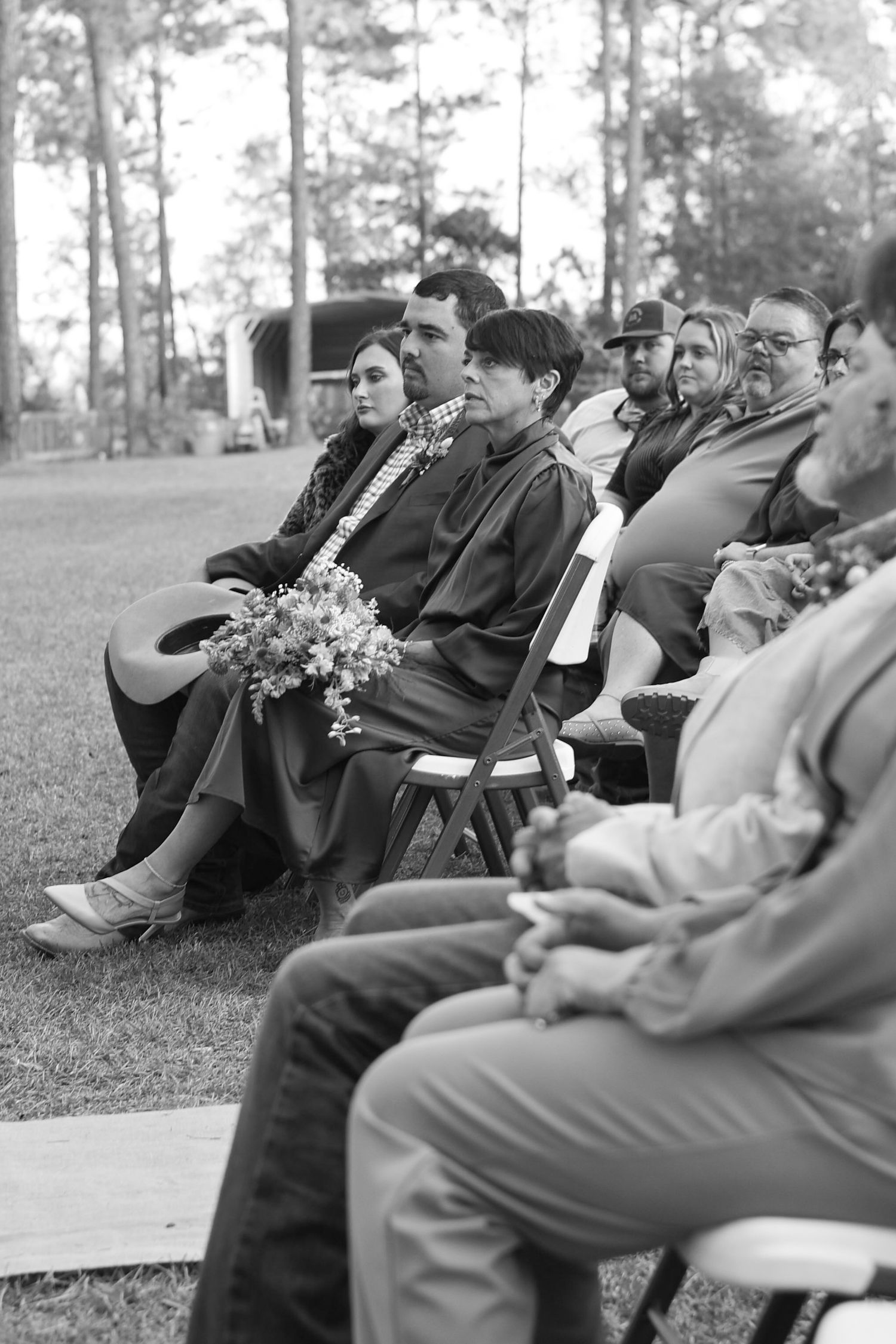 Wedding guests seated on white chairs in an outdoor pine forest setting during a ceremony.