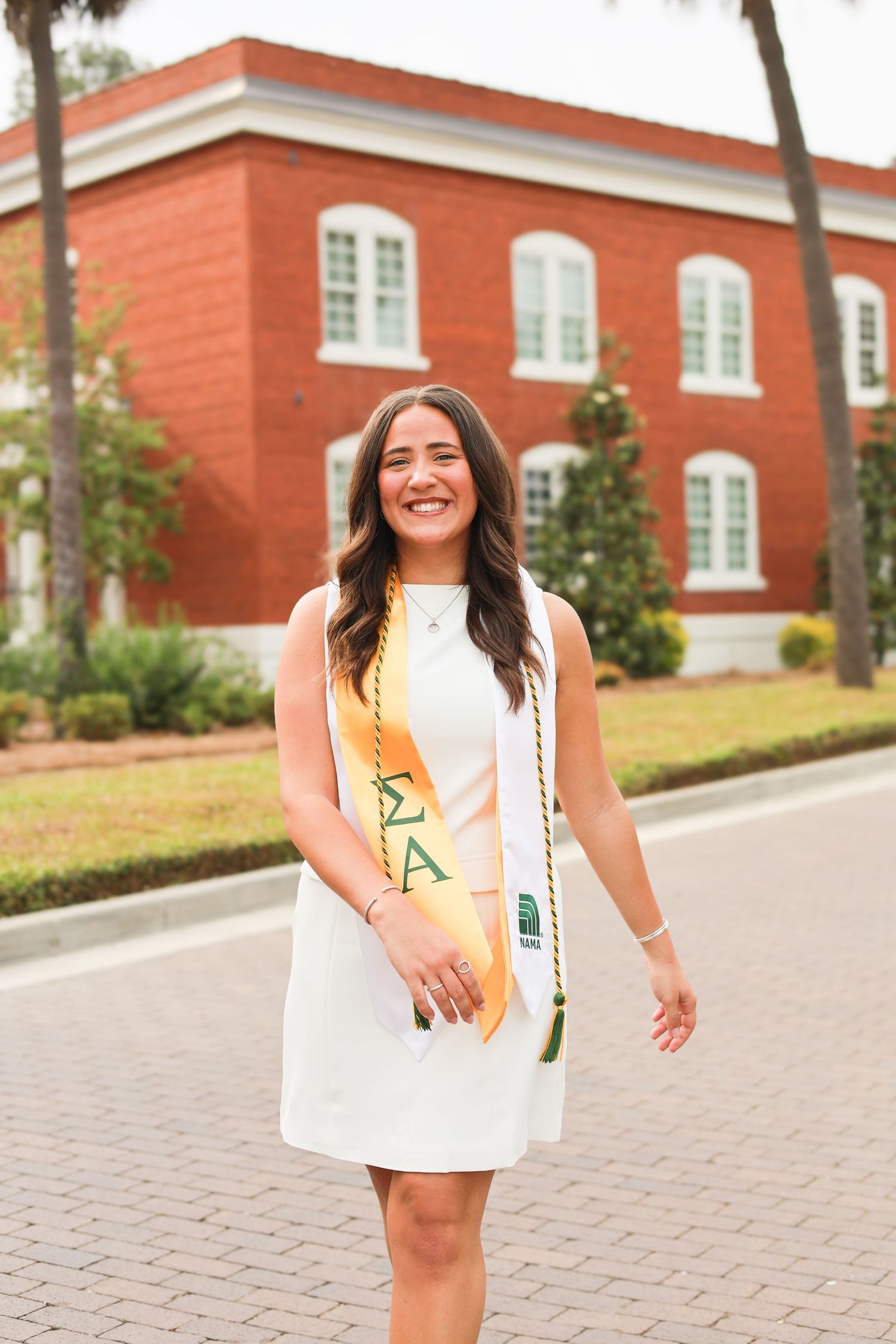 Graduate in white dress with sorority sash poses in front of red brick building.