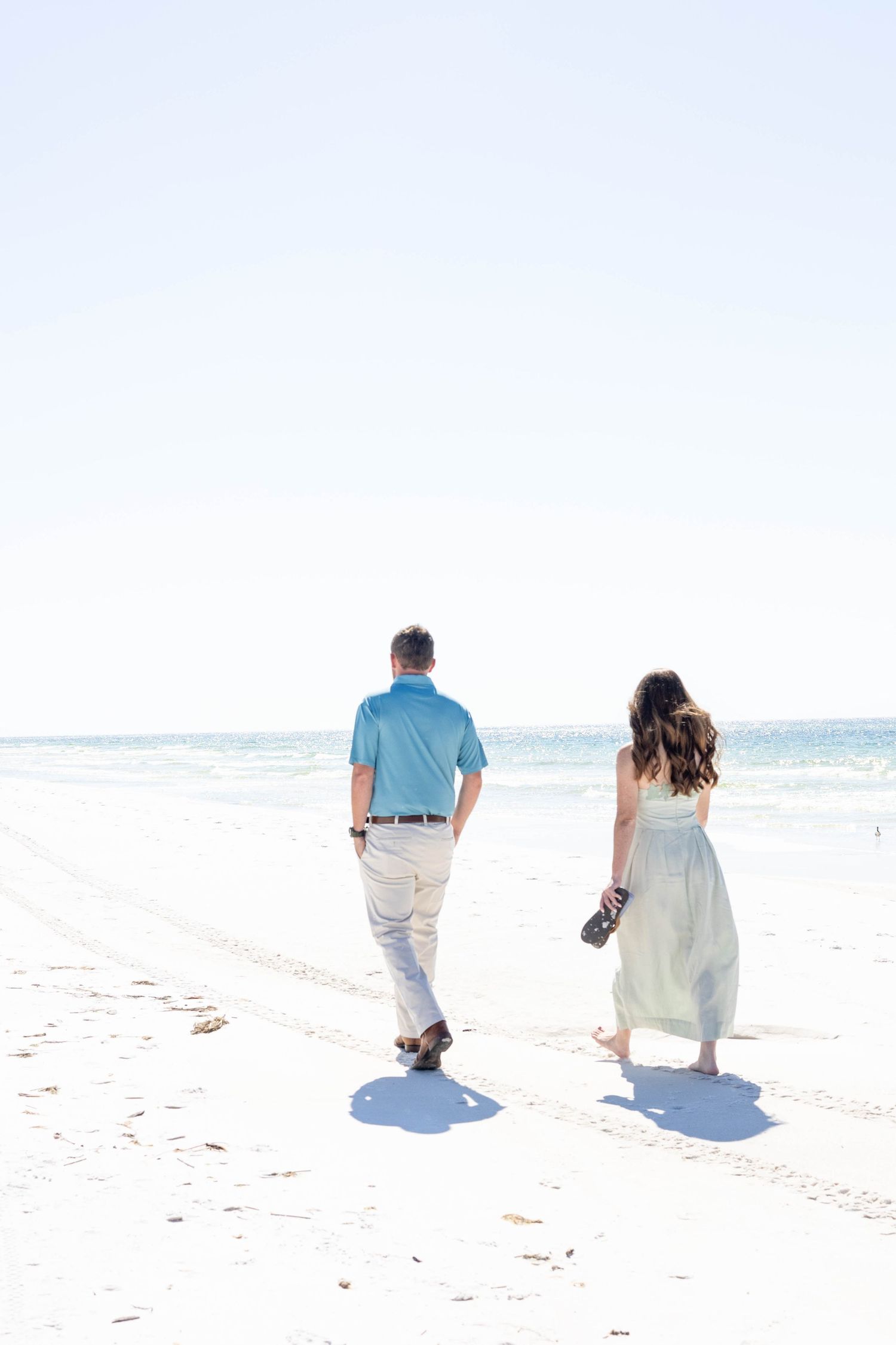 Couple walking hand in hand on a white sandy beach toward the ocean, woman in flowing dress carrying shoes.